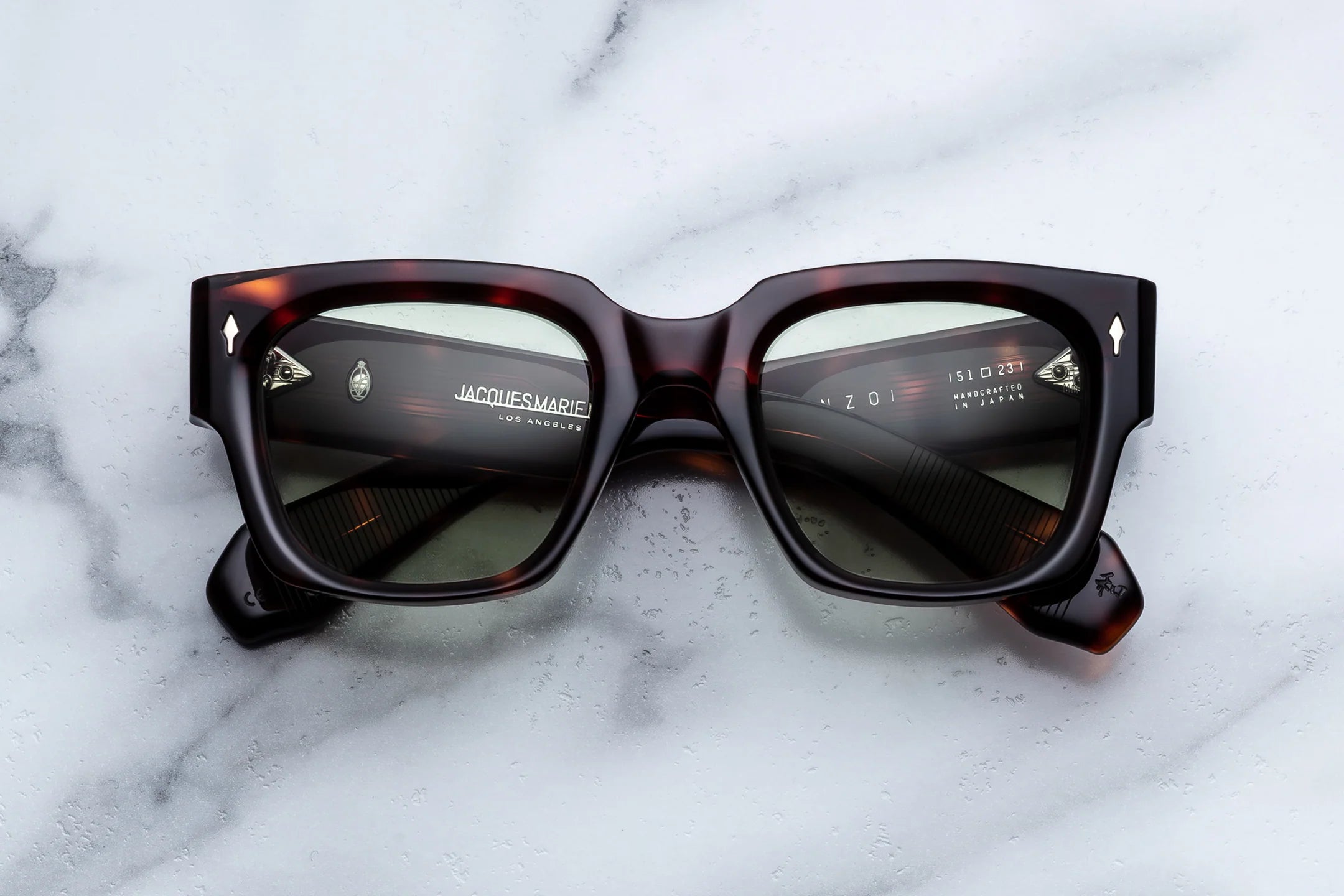 A pair of oversized square sunglasses with dark brown and amber tortoiseshell frames and dark lenses, resting on a white marble surface. The brand name 'JACOBSMART' is visible on the inner left temple arm.