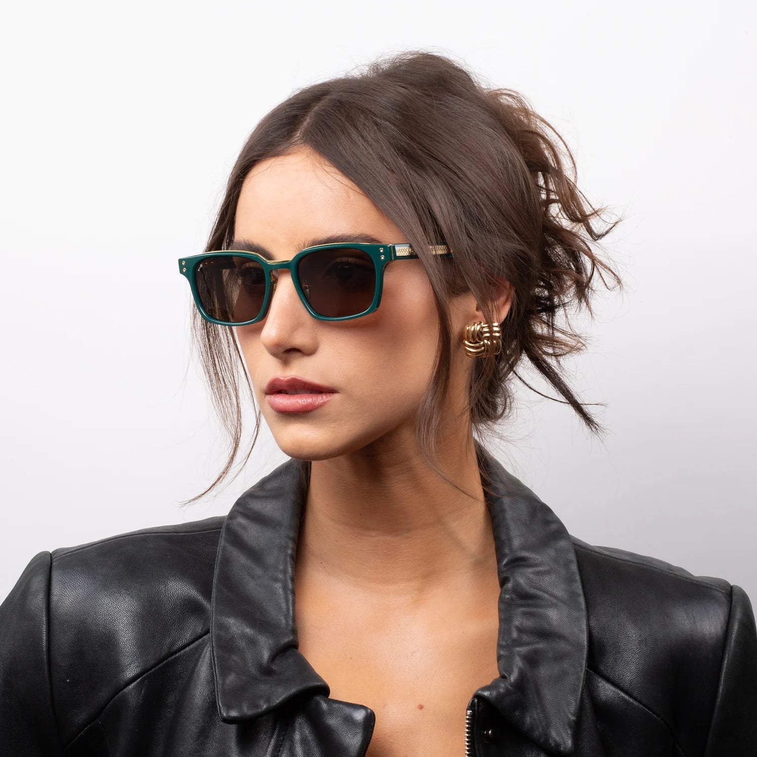 A woman with dark hair in a messy bun wearing green-framed sunglasses with dark lenses and a black leather jacket, photographed against a light gray studio background.