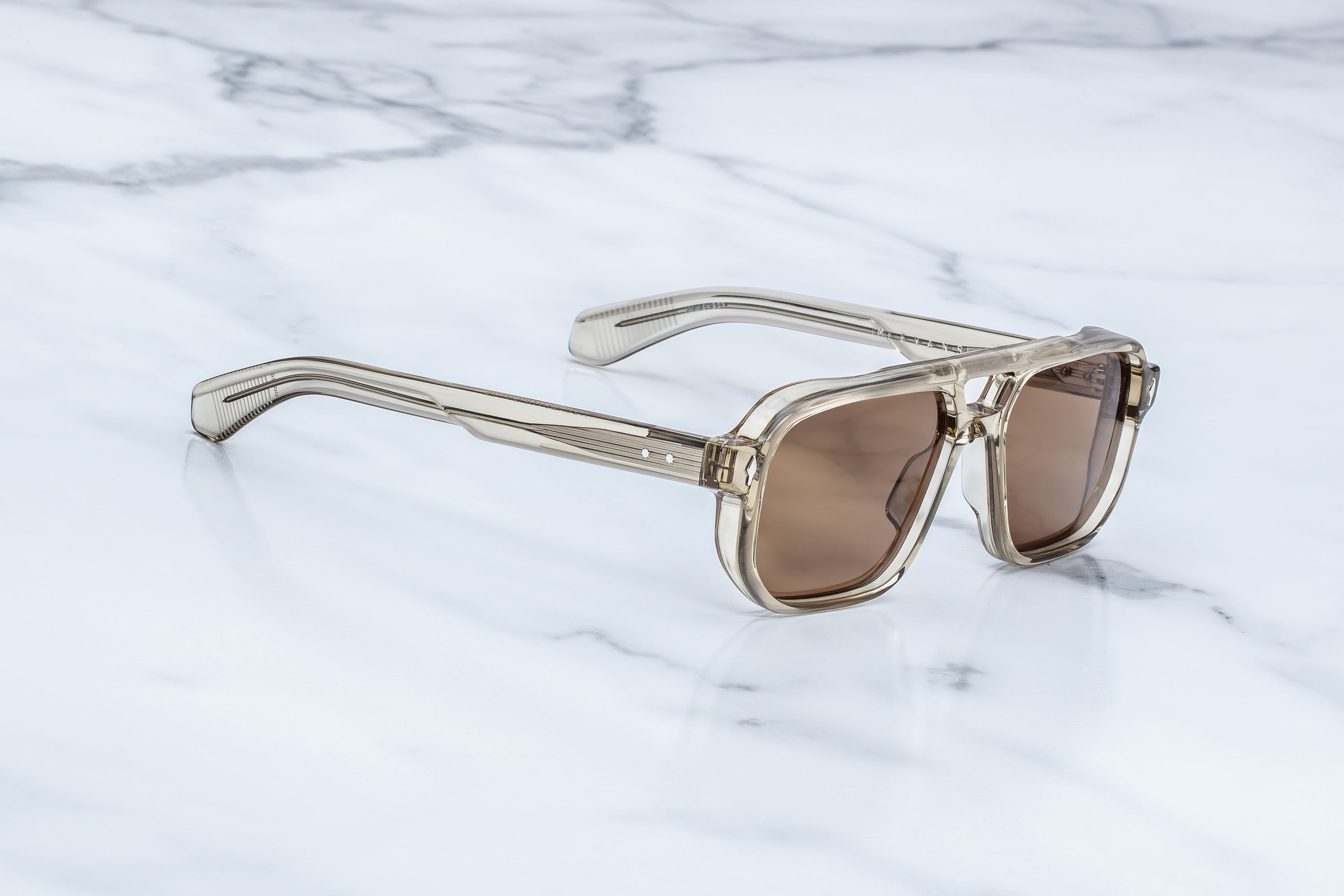 Translucent taupe acetate sunglasses with warm brown lenses, shown from an angled side view on a light marble background.