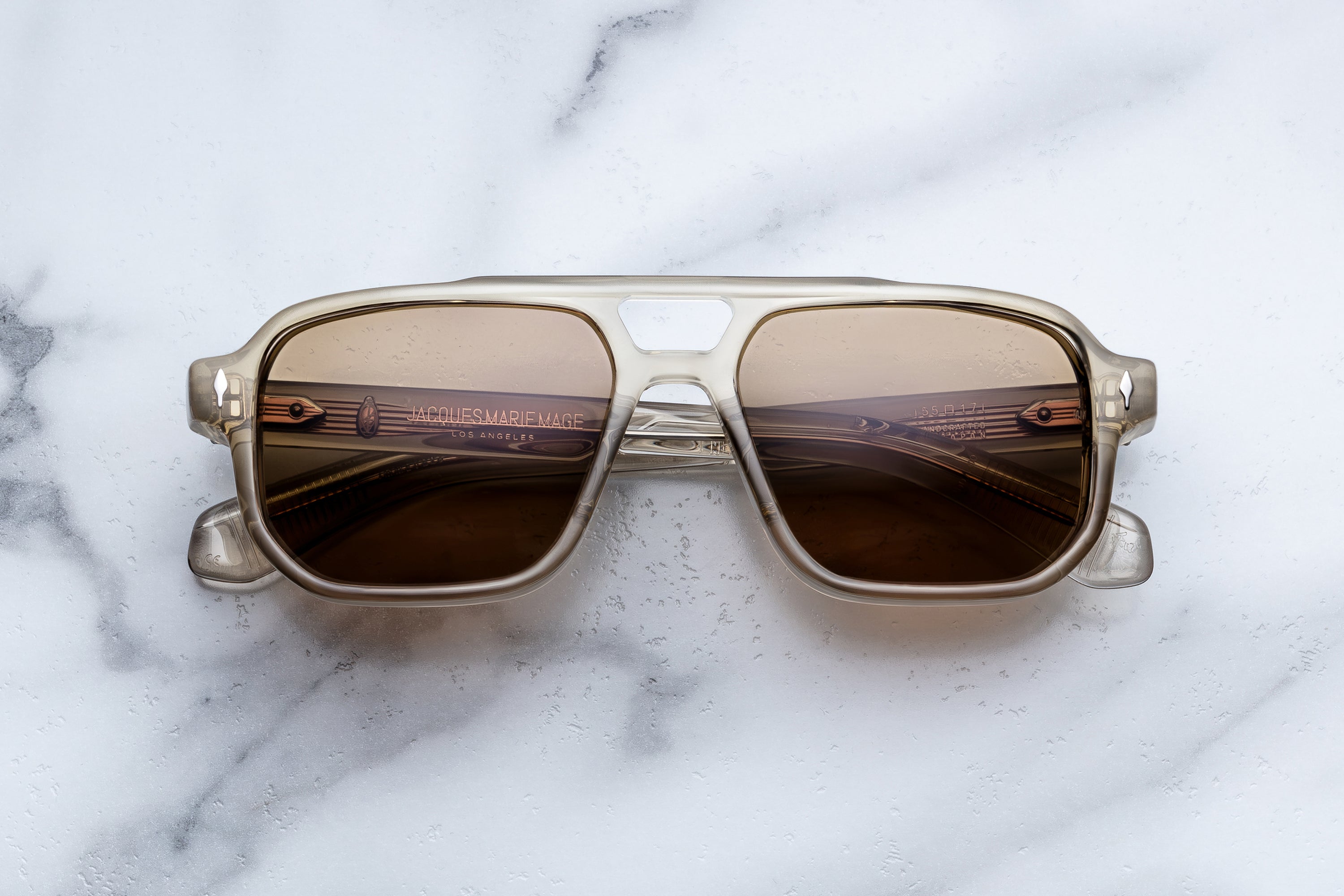 Jacques Marie Mage sunglasses with a translucent taupe acetate frame and warm brown lenses, shown front-facing on a light marble background.