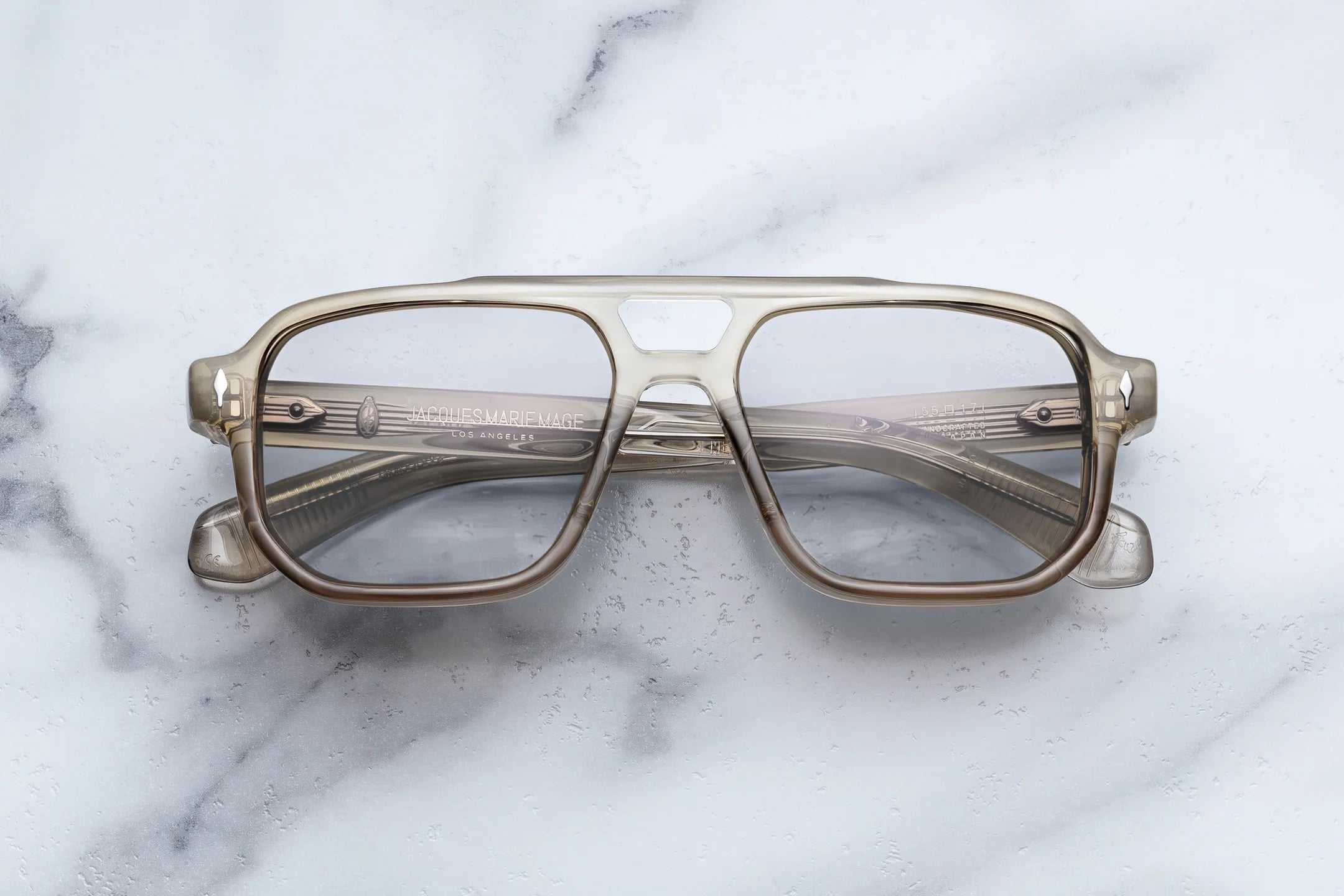 A pair of oversized square aviator-style eyeglasses with a translucent smoky gray acetate frame and clear lenses, resting on a white marble surface.