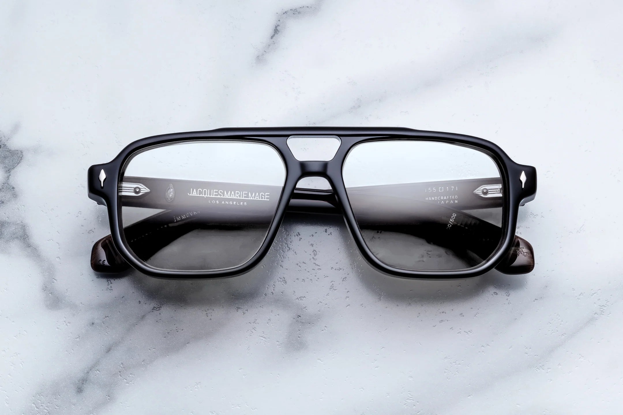 A pair of square-frame eyeglasses with a glossy black frame and dark gray lenses, resting on a white marble surface. 