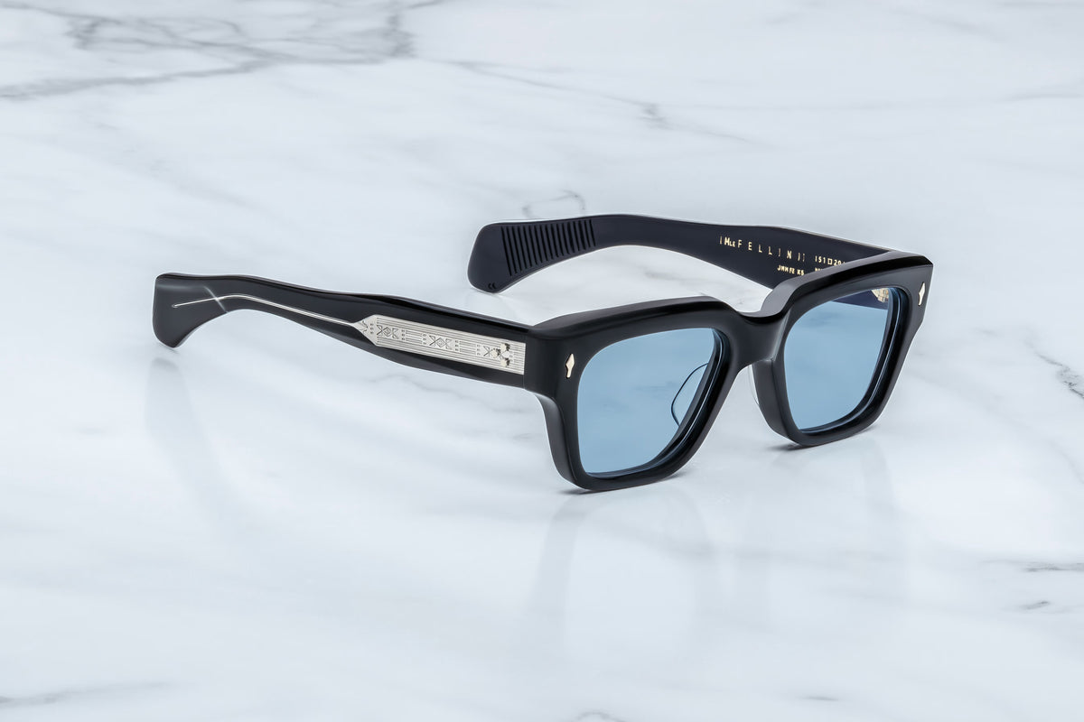 The Jacques Marie Mage Fellini by Jacques Marie Mage features black rectangular frames with light blue lenses, silver decorative accents along the arms, and angled temples inspired by iconic filmmaker styles, showcased elegantly on a white marble surface.