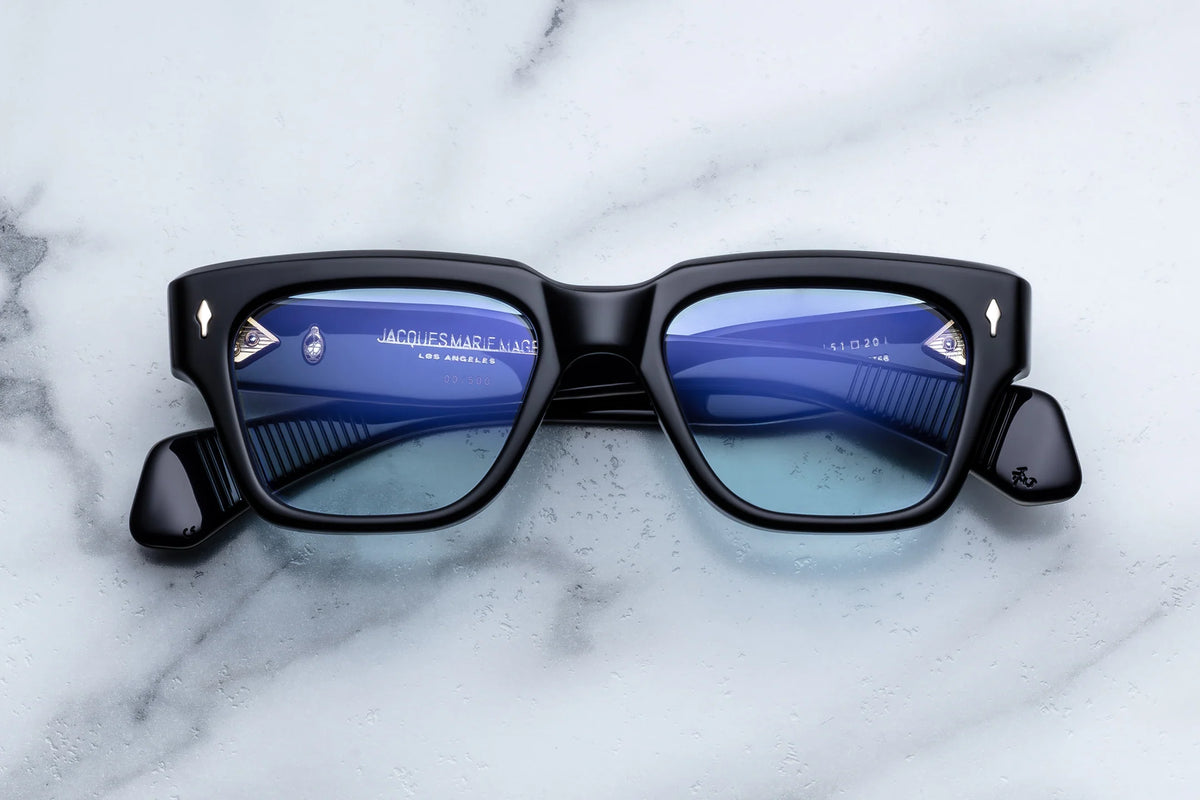 Jacques Marie Mage Fellini rectangular eyeglasses by Jacques Marie Mage, featuring black frames and blue-tinted lenses inspired by classic filmmakers, are displayed on a white marble surface. The reflection in the lenses reveals text and part of the inner arms.