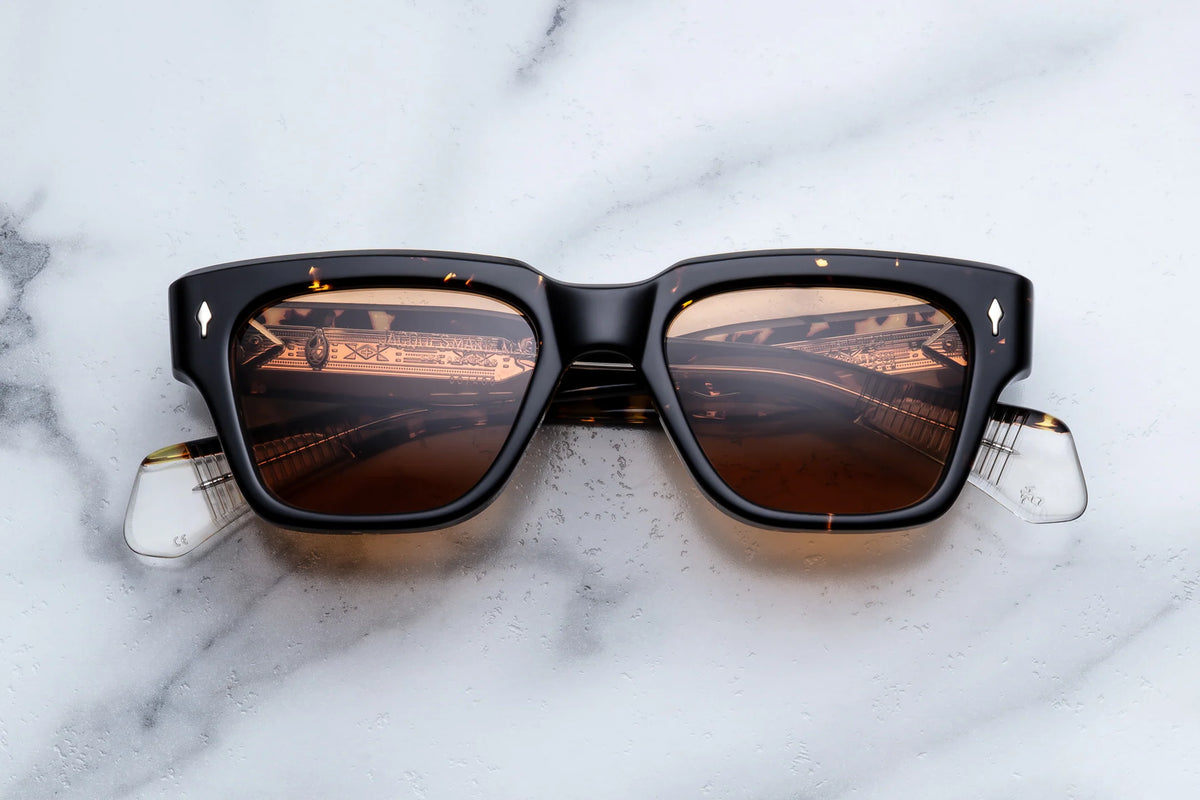 A pair of Jacques Marie Mage Fellini sunglasses by Jacques Marie Mage, featuring brown-tinted lenses and a sleek black frame, rest on a white marble surface. Reflections of a decorative ceiling or ornate interior are visible in the lenses, further enhancing the unique allure of this idiosyncratic eyewear.