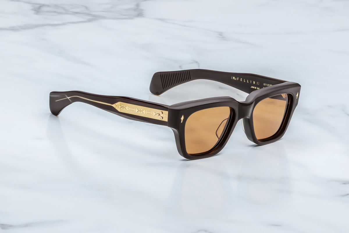 The Jacques Marie Mage Fellini sunglasses by Jacques Marie Mage feature a brown square frame with gold detailing on the temples, shown resting on a white marble surface. The lenses are tinted brown, and the inside of one arm displays engraved text—a subtle homage to classic spectacles, perfect for those inspired by filmmaker style.