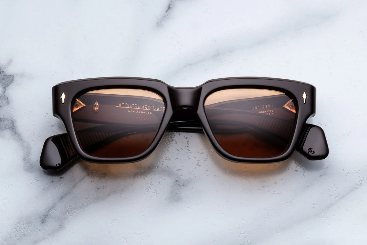Jacques Marie Mage Fellini sunglasses by Jacques Marie Mage, featuring brown rectangular frames with a thick, glossy finish, rest on a white marble surface. The distinctive eyewear showcases tinted lenses that faintly reflect what appears to be text from the inside of the arms.