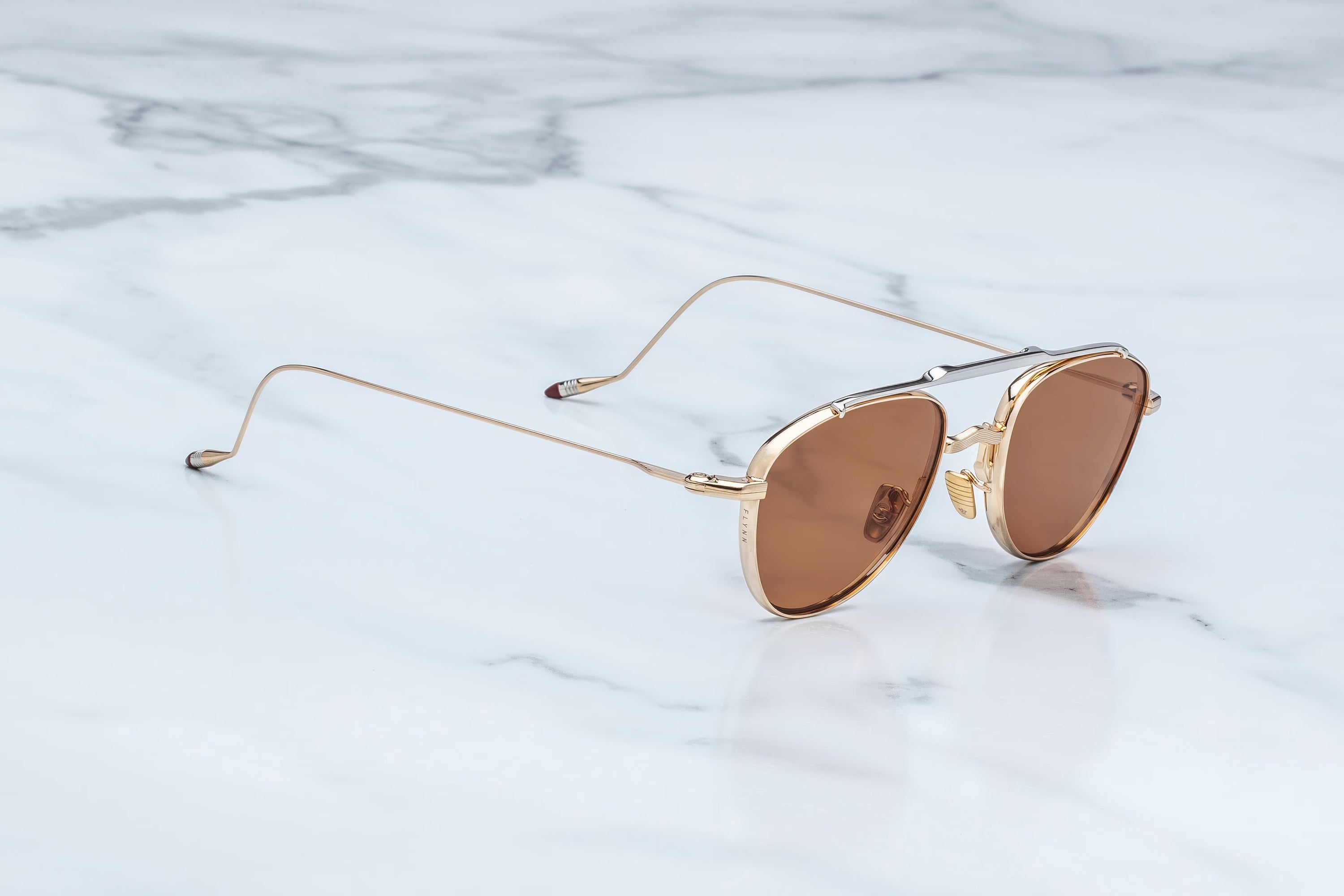 A pair of aviator-style sunglasses with gold metal frames and warm brown lenses, resting on a white marble surface.