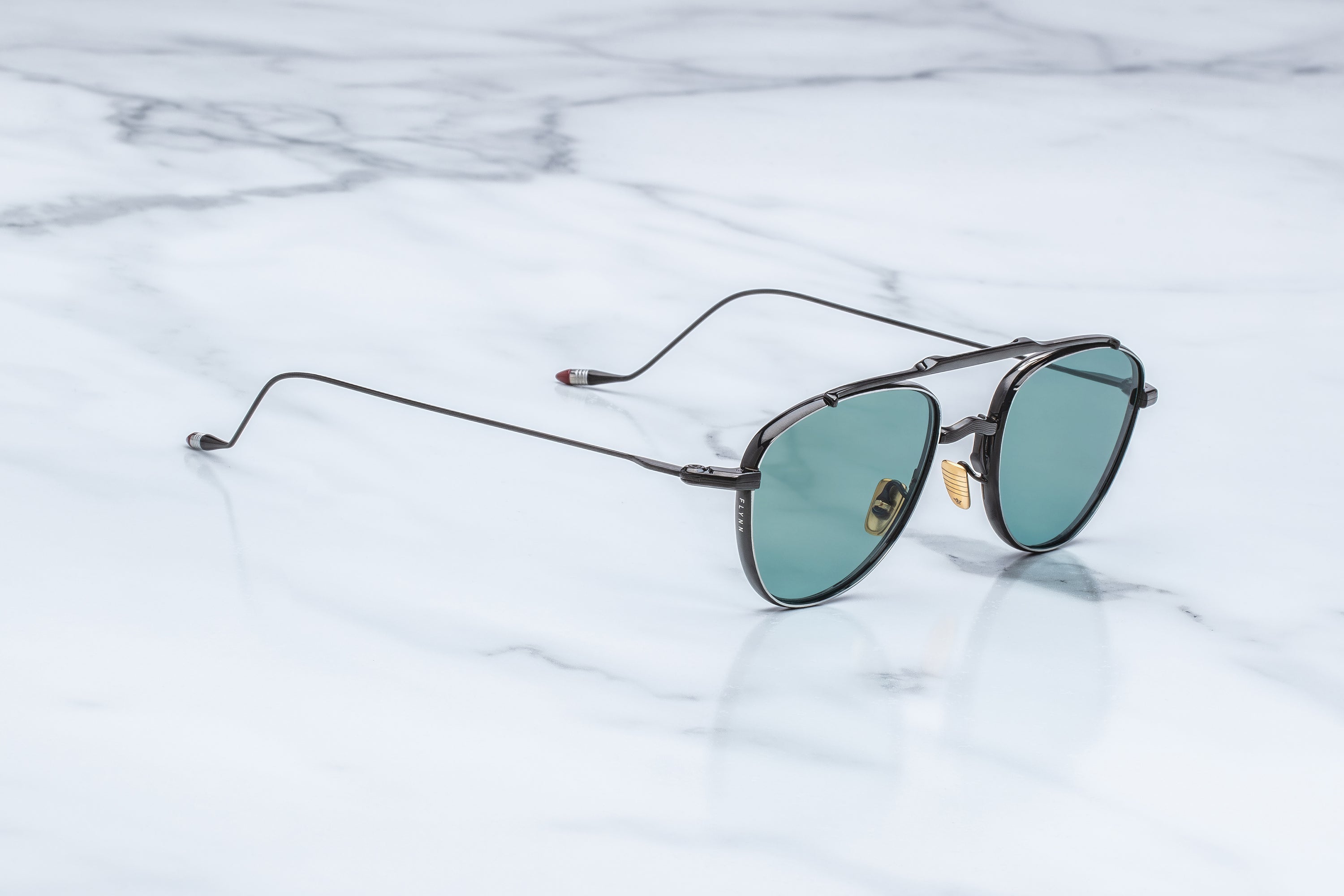 A pair of double-bridge aviator-style sunglasses with thin dark metal frames and green-tinted lenses, resting on a white marble surface.