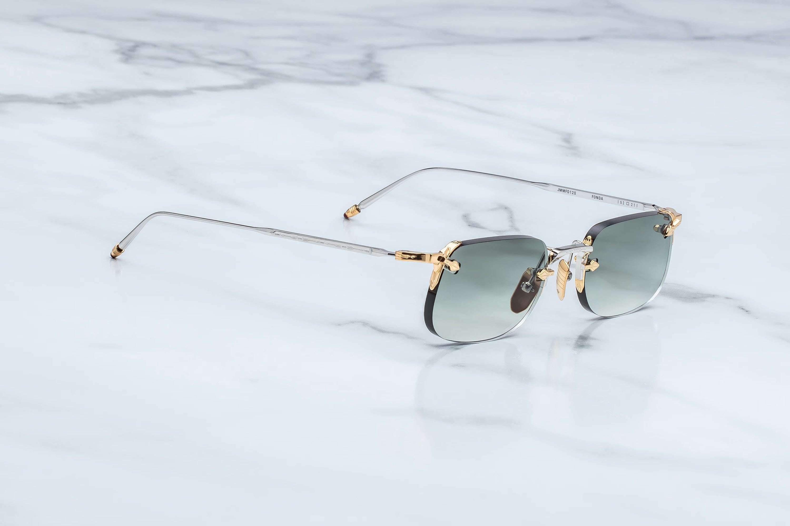 Rimless sunglasses with a thin silver-tone metal frame, gold-toned hinges, and dark gray gradient lenses, resting on a white marble surface.