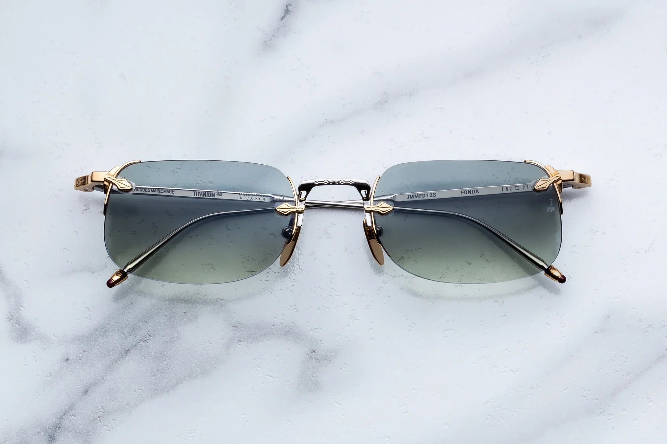 Rimless sunglasses with a gold-tone metal frame and gradient gray-green lenses, displayed on a white marble surface.