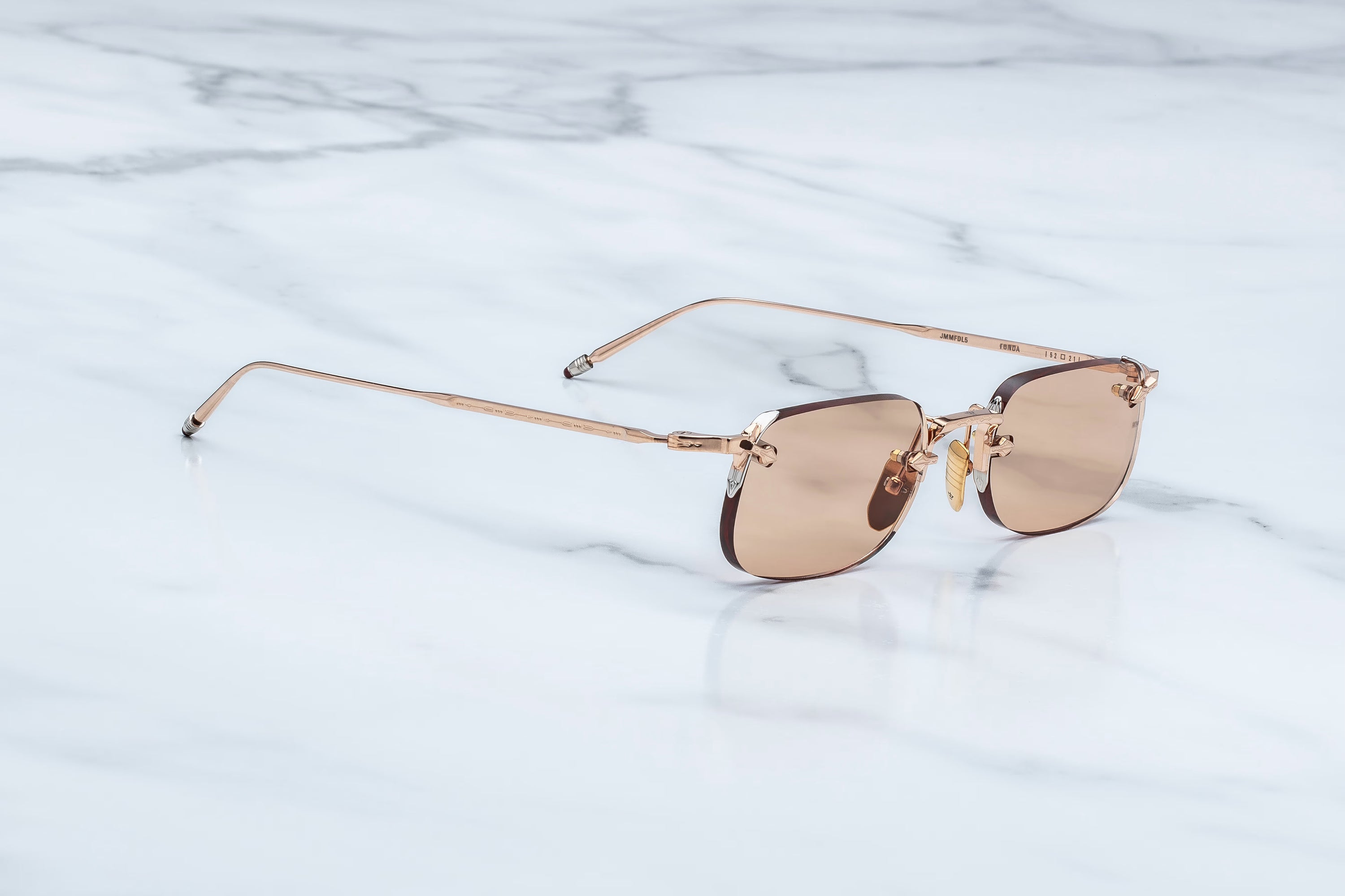 Rimless sunglasses with a thin rose gold metal frame and warm brown tinted lenses, resting on a white marble surface.