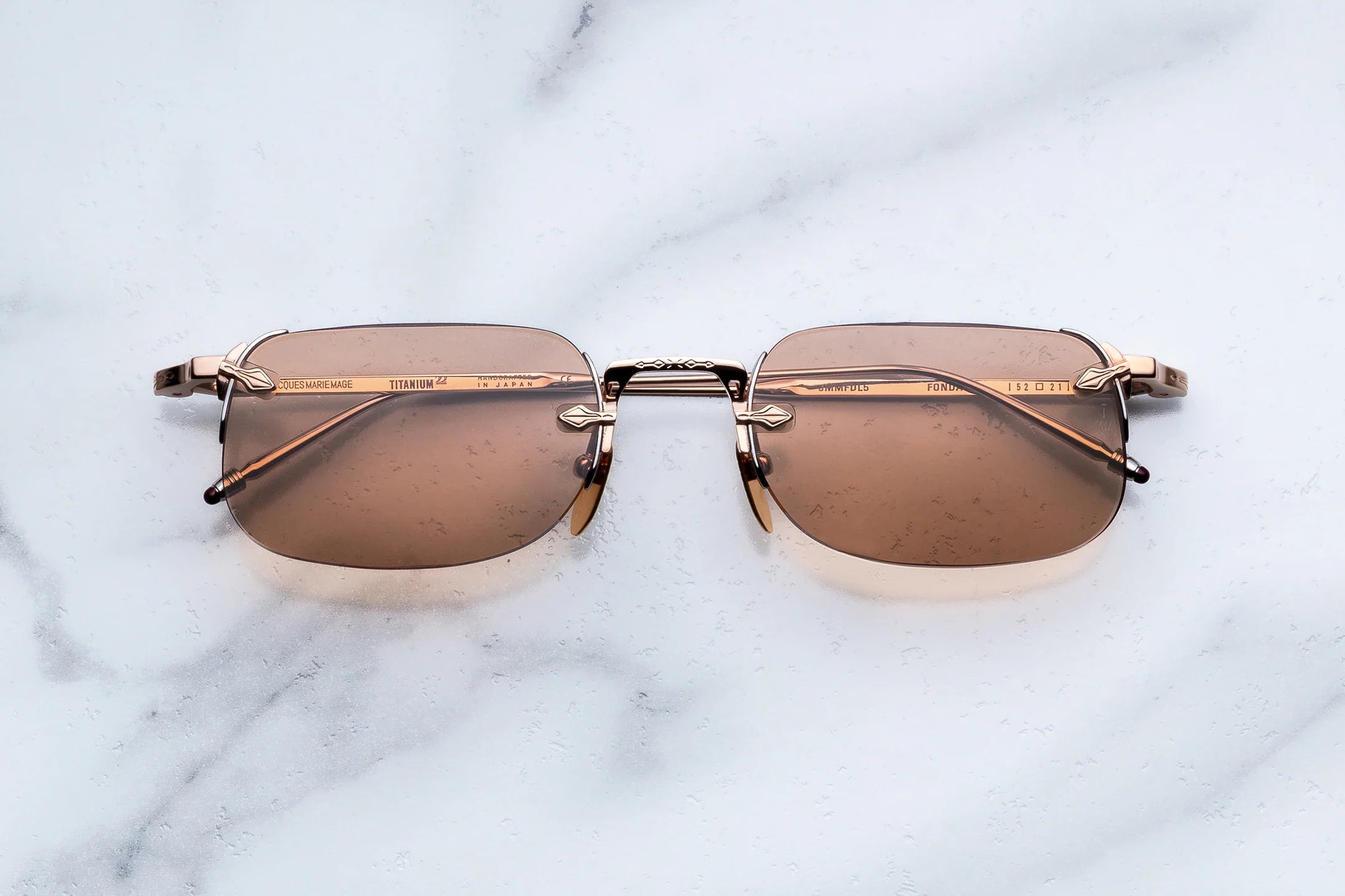 Rimless square sunglasses with brown tinted lenses and gold metal frames, displayed on a white marble surface.