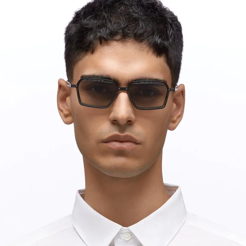 A man wearing black square-frame sunglasses with dark tinted lenses, shown from the chest up against a plain white background.
