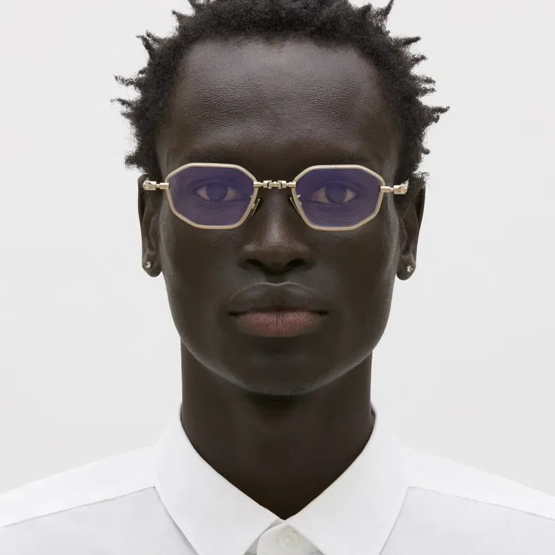 A close-up portrait of a model wearing gold-framed sunglasses with purple tinted lenses and a white collared shirt against a light gray background.