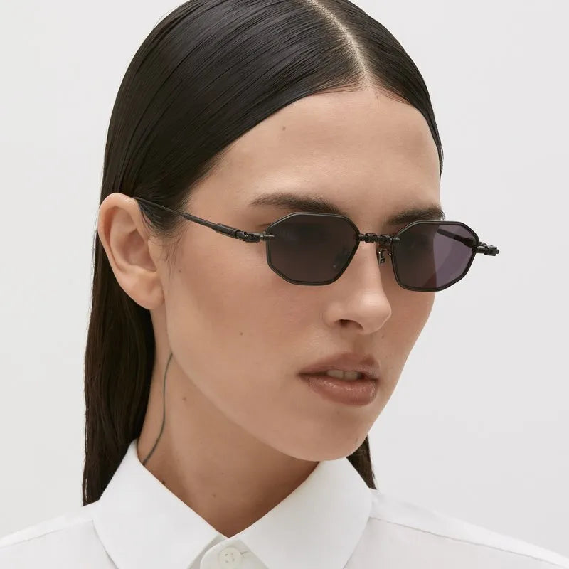 A person with straight, dark hair, wearing the Kuboraum H66 small black designer sunglasses and a white collared shirt, looks slightly to the side. A thin tattoo line is visible on the left side of their neck, reflecting the bold spirit of Kuboraum’s eyewear.