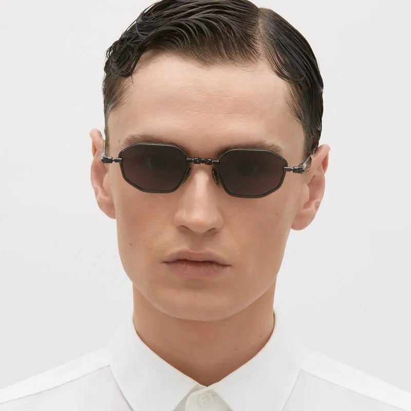 A person with short, neatly combed hair wears the Kuboraum H66 small, oval black sunglasses by Kuboraum—handmade in Italy—and a white buttoned shirt, looking straight at the camera against a plain background.