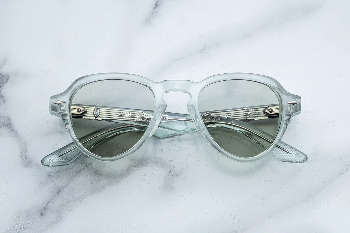Jacques Marie Mage Hatfield clear-frame glasses with light grey photochromic lenses resting on a white marble surface.