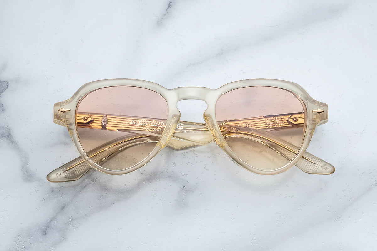 Jacques Marie Mage Hatfield sunglasses in translucent beige with a round frame, gradient peach photochromic lenses, and exposed metal core wire inside the temples.