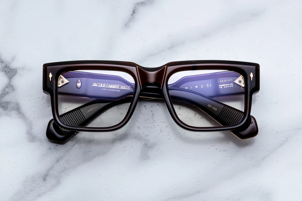 Dark brown rectangular Jacques Marie Mage Hemmings eyeglasses with gold arrow-shaped hinges resting on a marble surface, channeling 60s-inspired spectacles.
