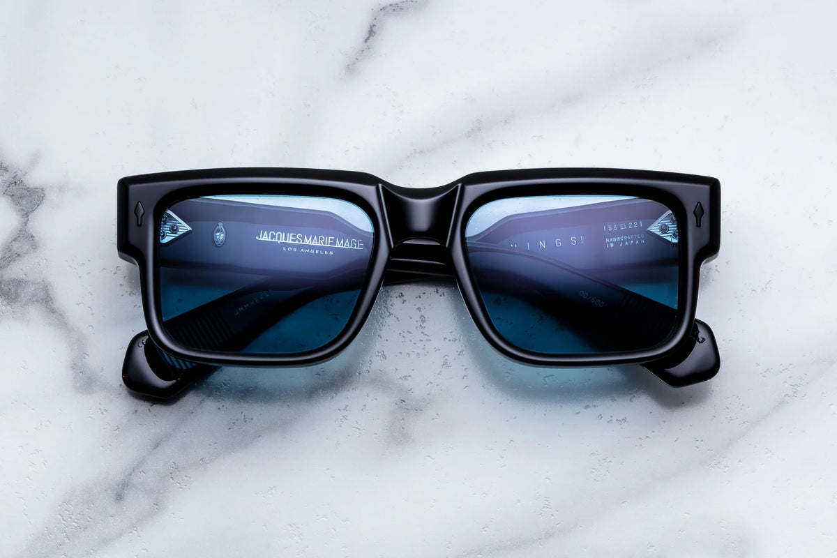 Jacques Marie Mage Hemmings 60s-inspired black rectangular sunglasses with blue-tinted lenses resting on a marble surface.