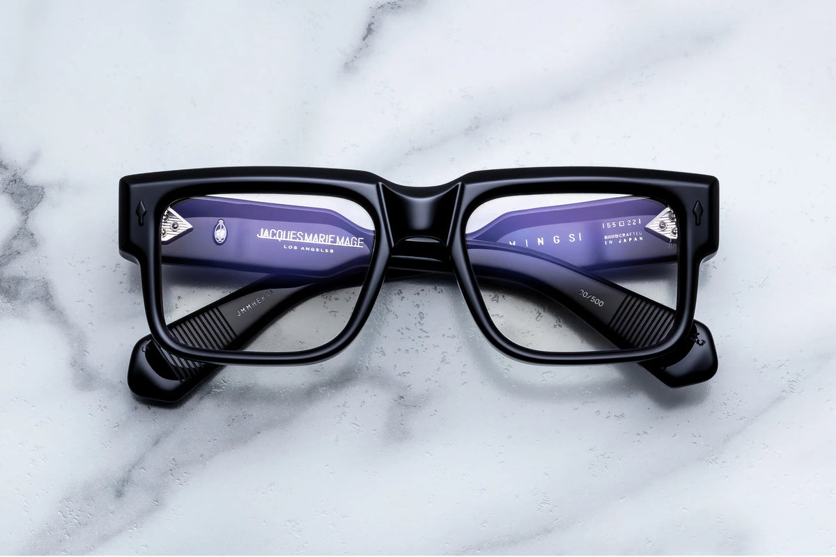 Jacques Marie Mage Hemmings sophisticated black rectangular eyeglasses with thick frames and silver temple accents, resting on a white marble surface in a 60s-inspired style.