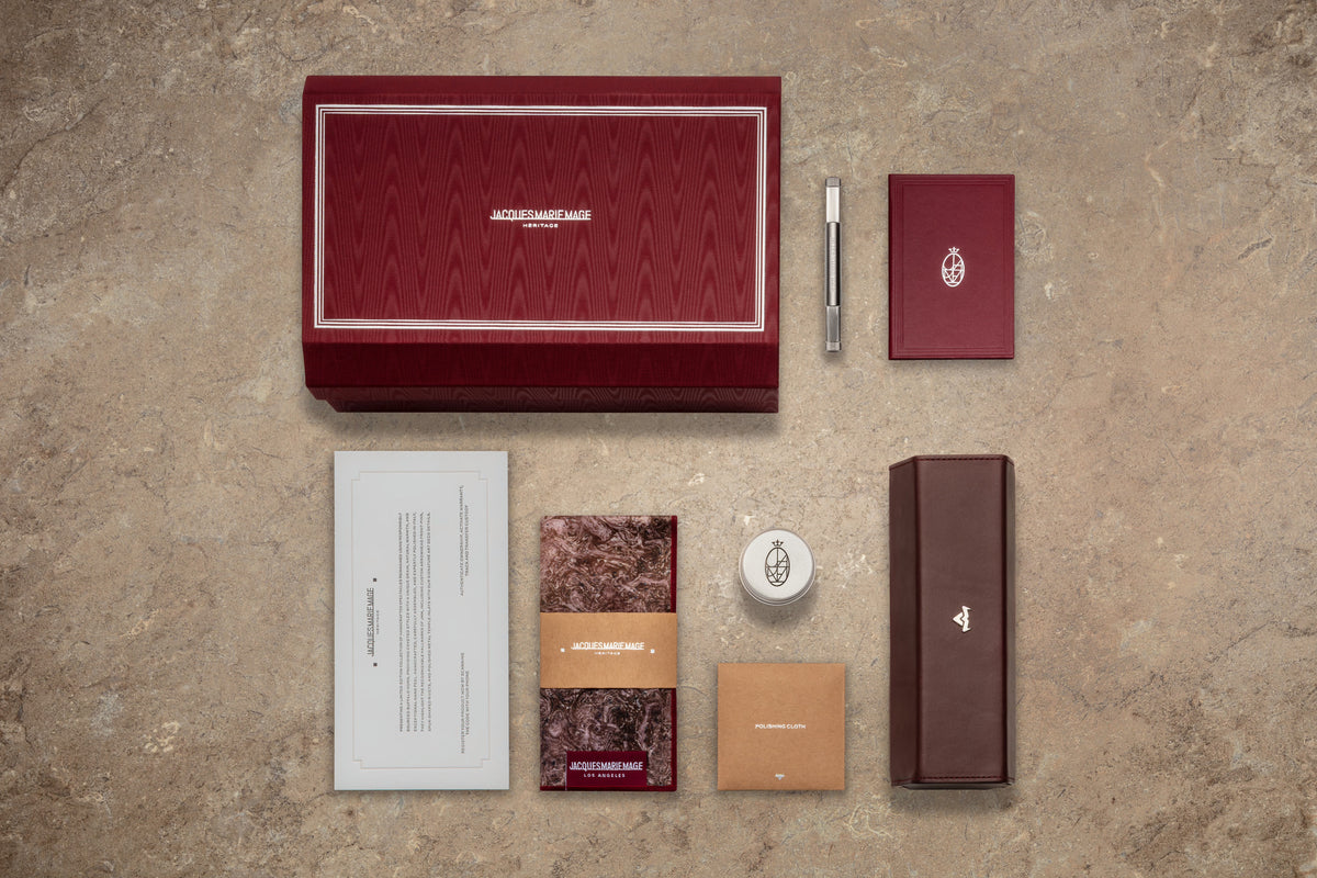 A luxury gift set on a beige surface features a red patterned box, pen, two smaller boxes, a candle, an envelope, and a booklet—all arranged elegantly. Completing the sophisticated display are exquisitely crafted Jacques Marie Mage Heritage 2: Leclair spectacles by Jacques Marie Mage, seamlessly blending timeless style with modern sophistication.