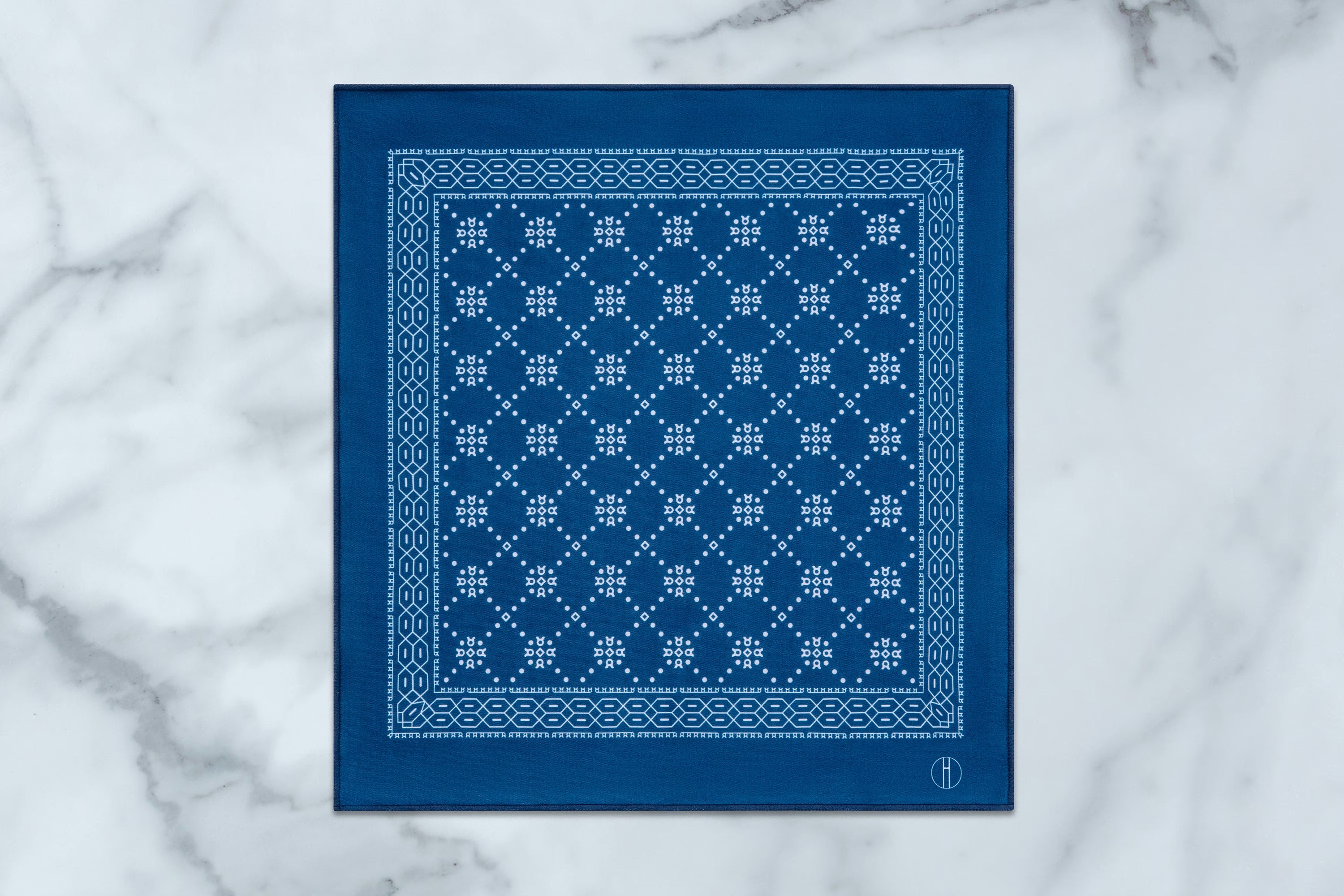 A square blue bandana with a white geometric lattice pattern and border, laid flat on a white marble surface.