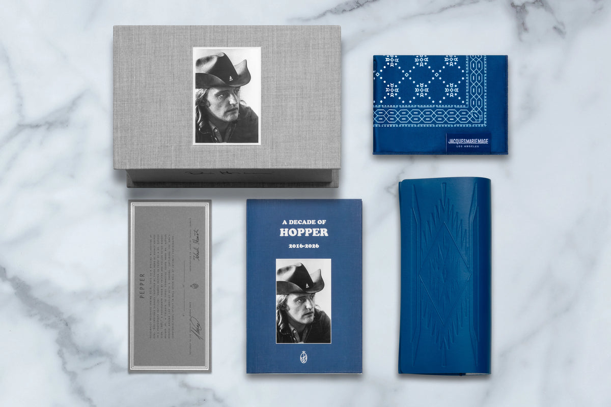 A gray box containing a Dennis Hopper collection portrait, a blue book, patterned cloth, and an information card displayed on a marble surface alongside Jacques Marie Mage A Decade of Hopper: Pepper eyewear.
