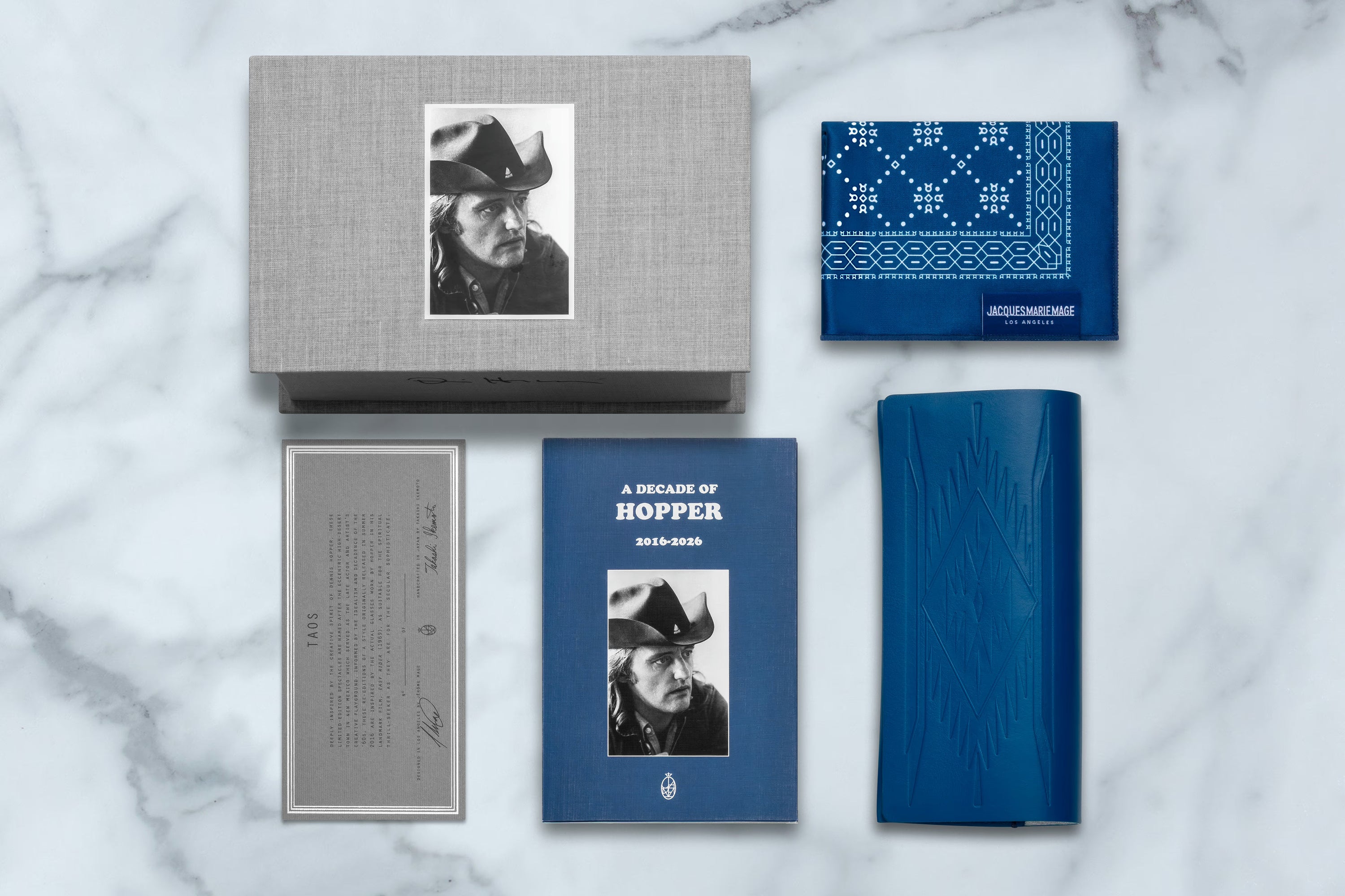 A curated flat lay of a book and matching merchandise on a white marble background. The set includes a blue hardcover book titled 'A Decade of Hopper' with a black-and-white portrait, a gray fabric-covered box with a matching portrait print, a blue bandana-style cloth with white geometric patterning, a gray printed card, and a blue textured sleeve or case.
