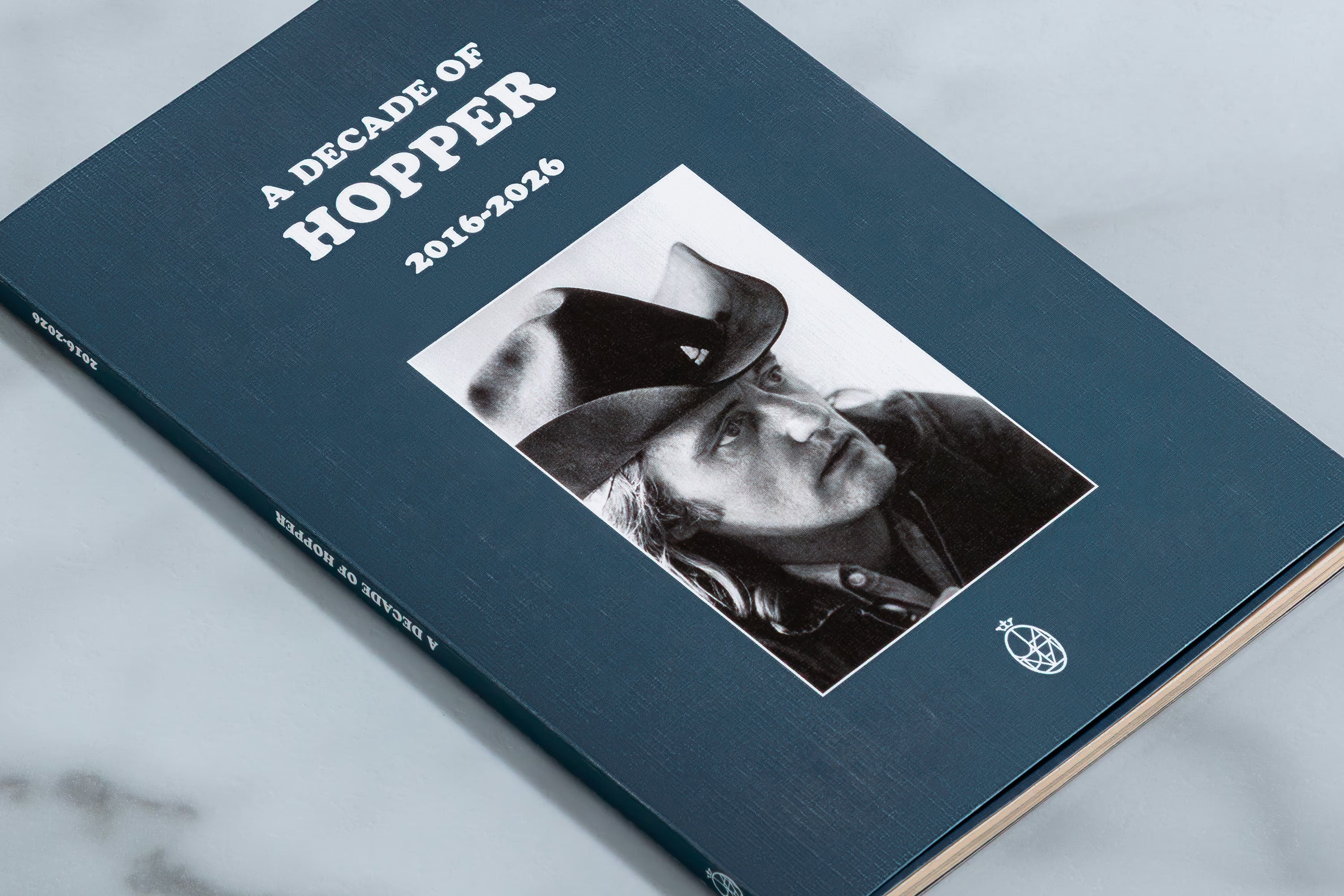 A hardcover book titled 'A DECADE OF HOPPER 2016-2026' resting on a light marble surface. The cover is a deep navy blue with white text and a black-and-white photograph of a cowboy in a hat.