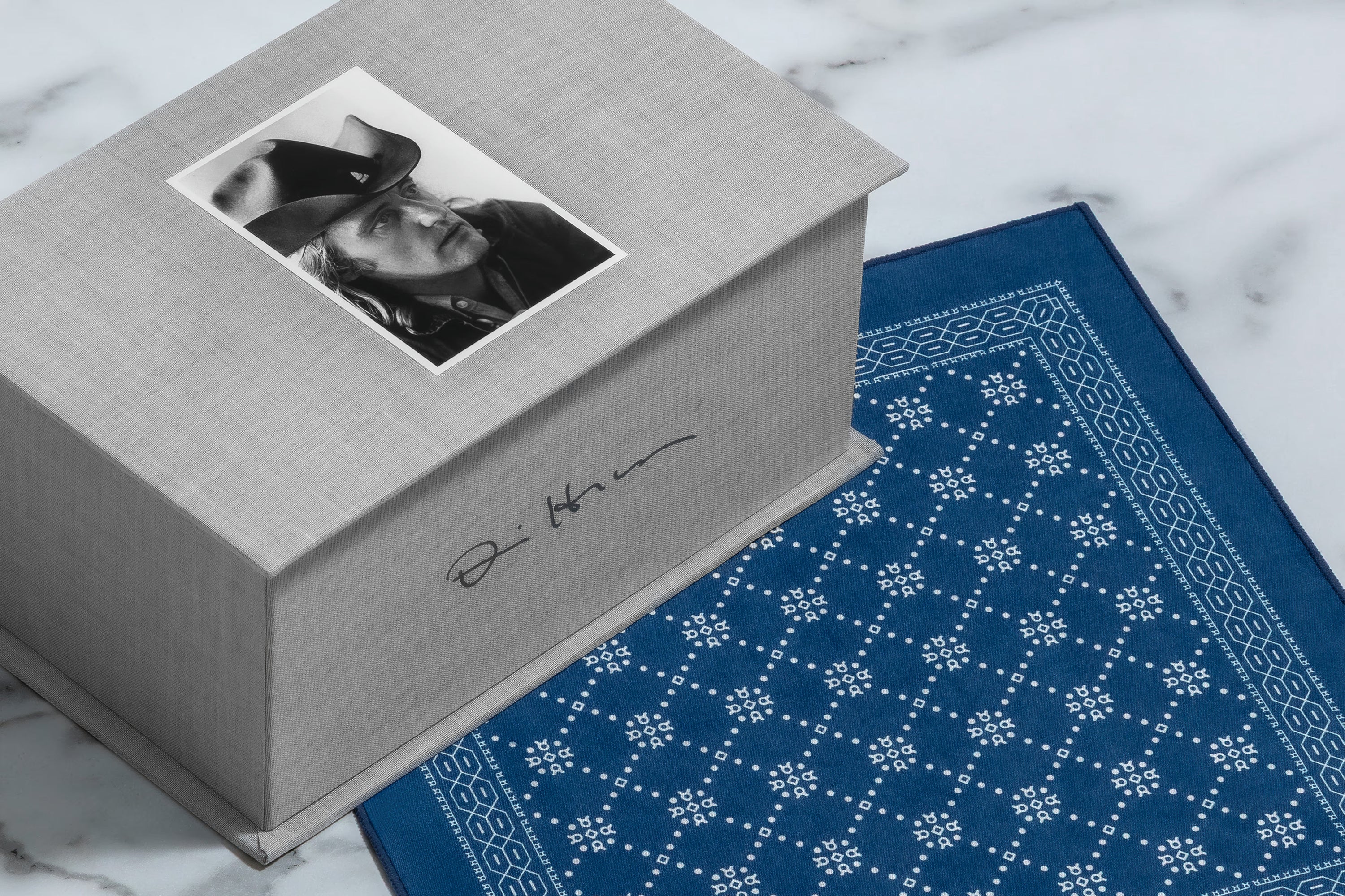 A gray fabric-covered presentation box with a black-and-white portrait photo on top and a signature on the side, placed on a white marble surface next to a blue patterned bandana.