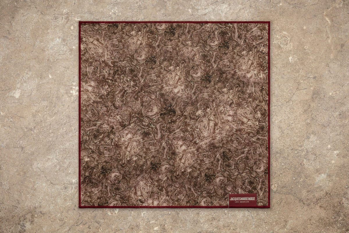 A square brown patterned handkerchief with marbled swirls, bordered in dark red, lies flat on a textured beige surface. In the corner, a small label is visible alongside the Jacques Marie Mage Heritage 2: Leclair eyewear by Jacques Marie Mage, perfectly completing this vintage-inspired scene.