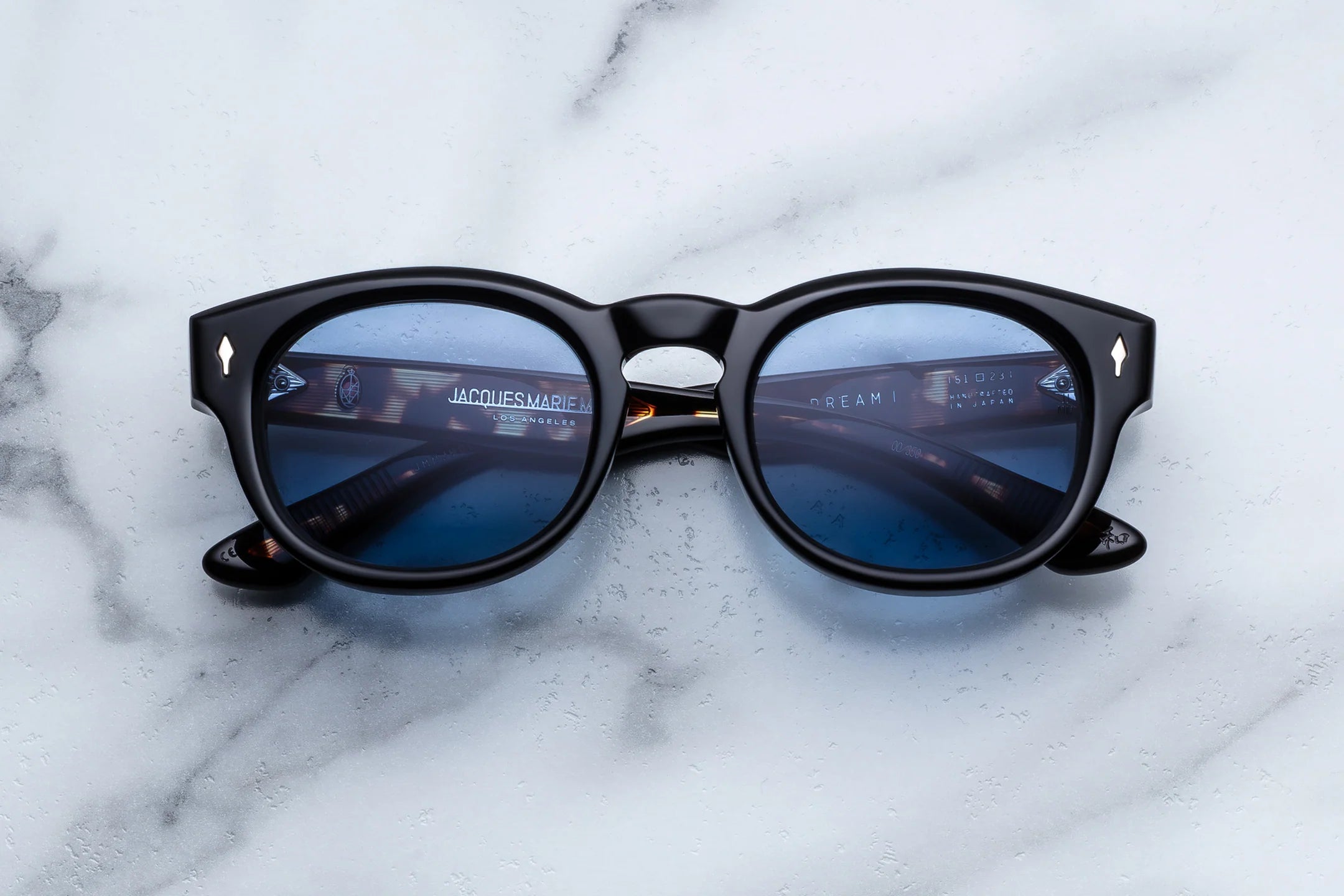 Jacques Marie Mage Chunky Black sunglasses with blue lenses on a marble surface