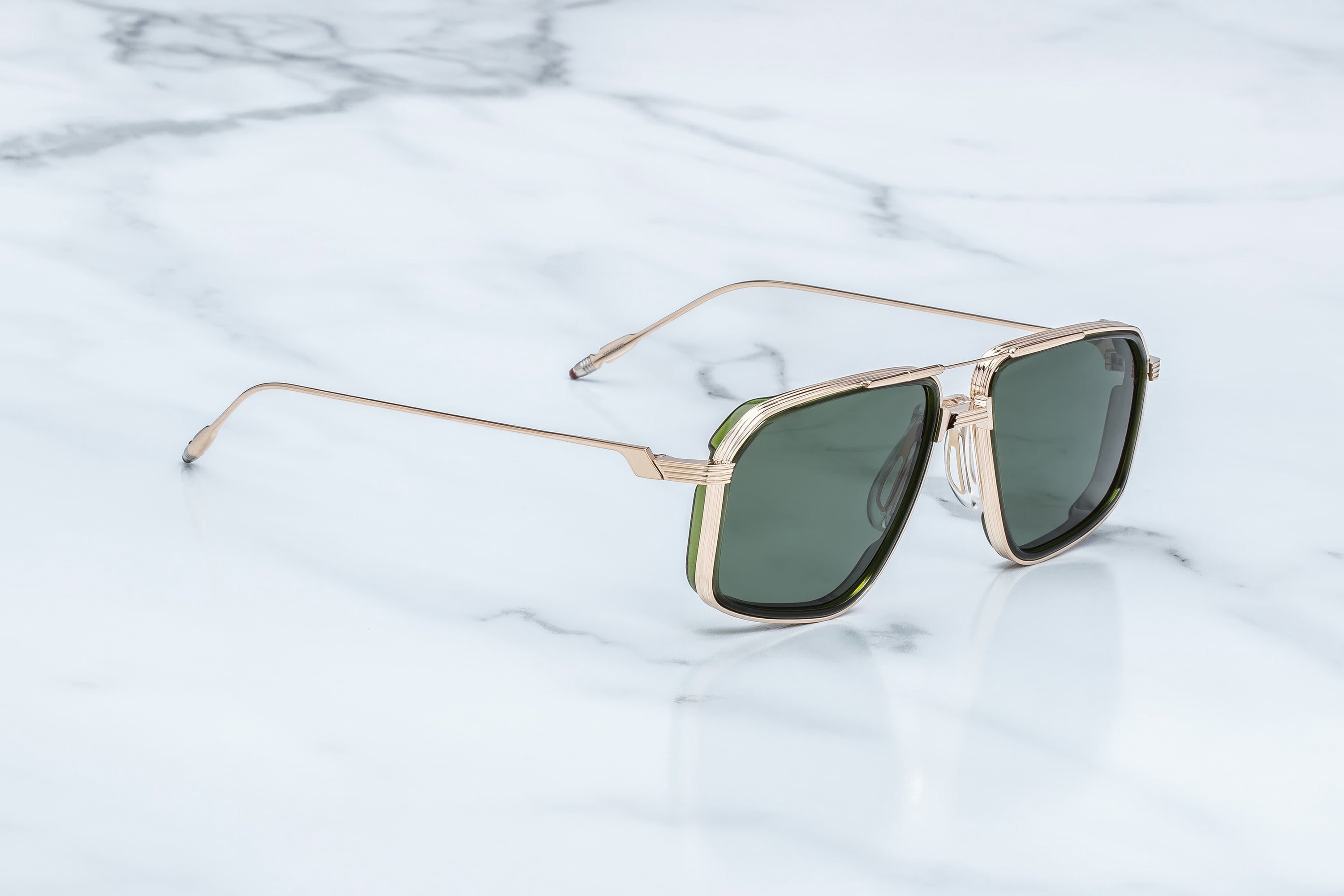 A pair of square aviator-style sunglasses with a gold metal frame and dark green lenses, resting on a white marble surface.