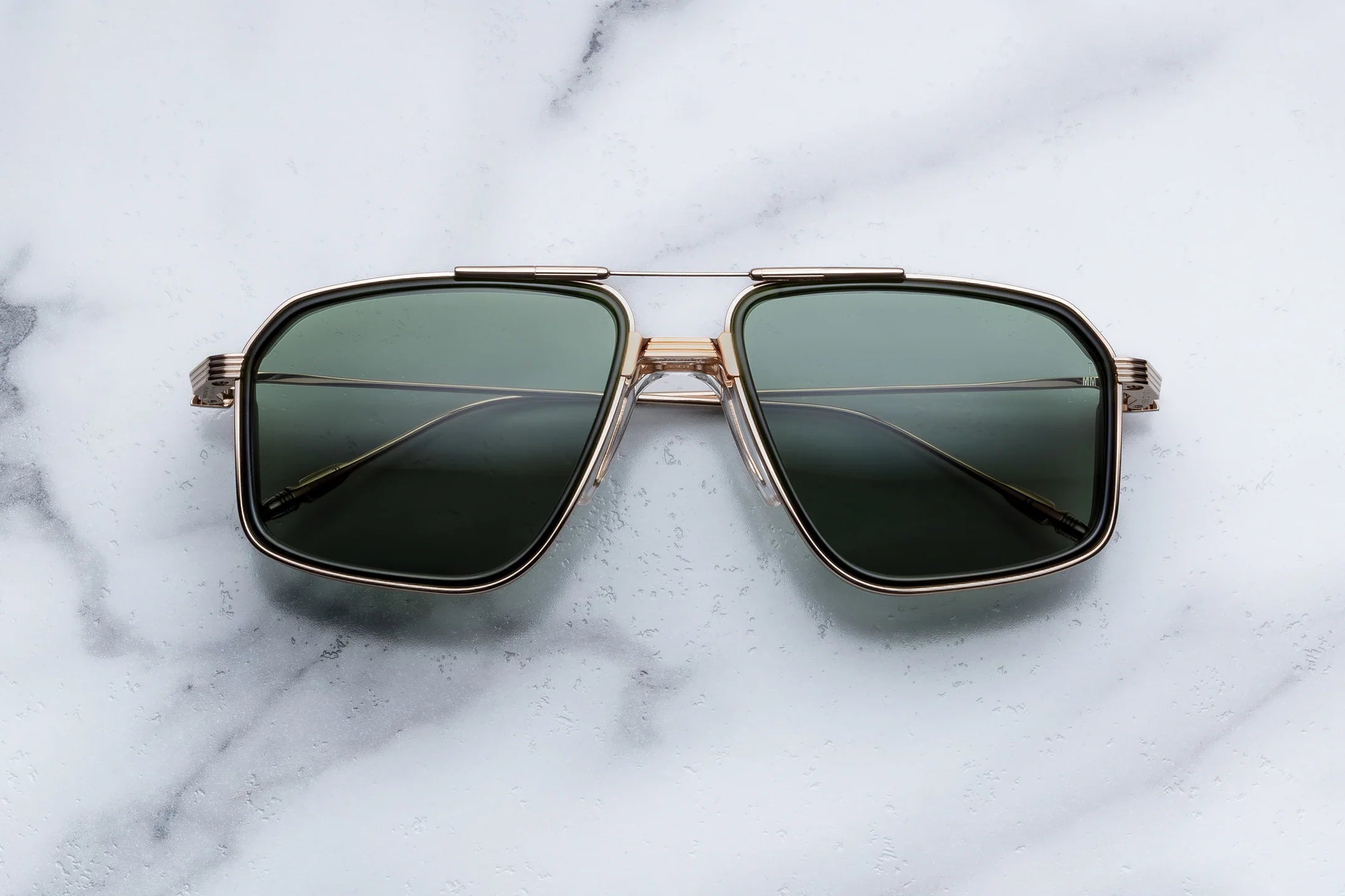 A pair of oversized square aviator-style sunglasses with dark green lenses and a gold-tone metal frame, resting on a white marble surface.