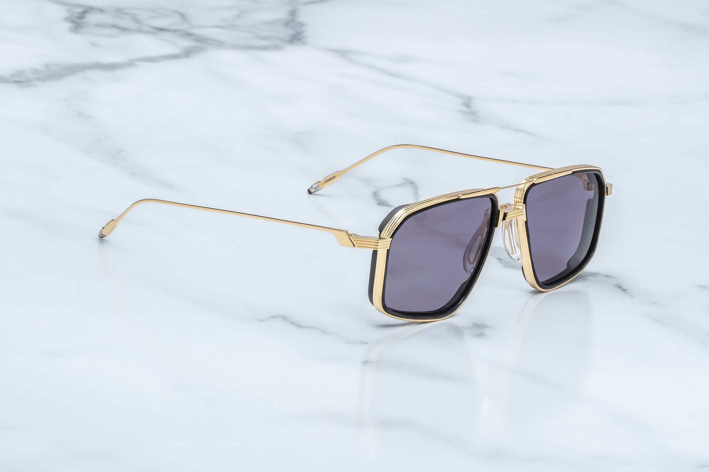 A pair of square aviator-style sunglasses with a gold metal frame and dark gray lenses, resting on a white marble surface.