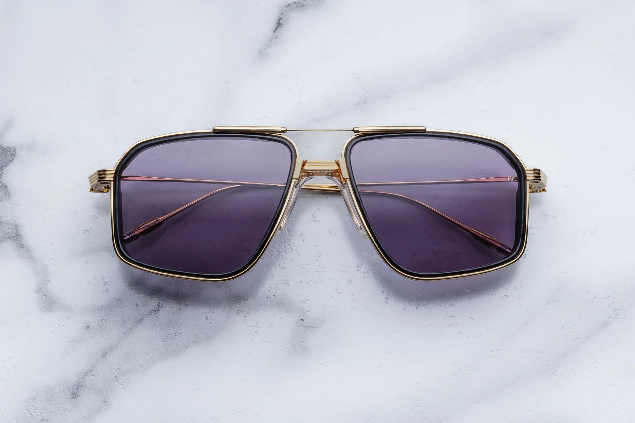 A pair of oversized square aviator-style sunglasses with a gold-tone metal frame and dark purple gradient lenses, displayed on a white marble surface.