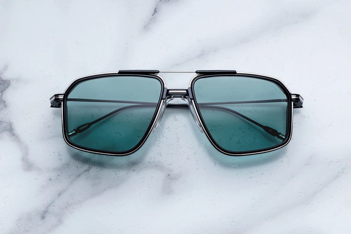 Jacques Marie Mage Jagger rectangular aviator sunglasses with black rims and teal-tinted lenses resting on a marble surface, capturing an air of animal magnetism.