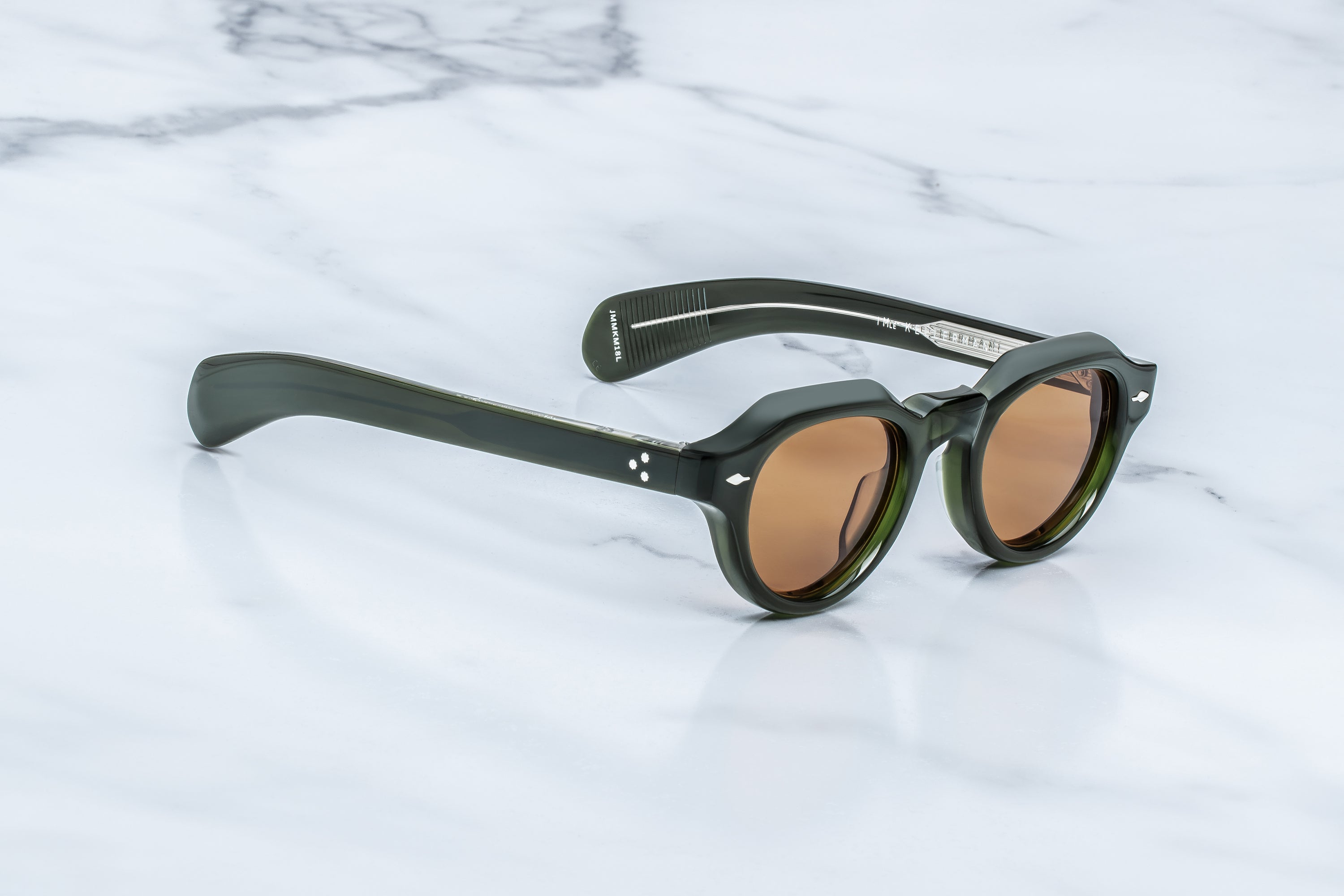 Jacques Marie Mage Kellerman sunglasses in dark green, featuring round designer frames with brown-tinted lenses, rest on a white marble surface. The thick-framed eyewear by Jacques Marie Mage displays small white markings near the hinges.
