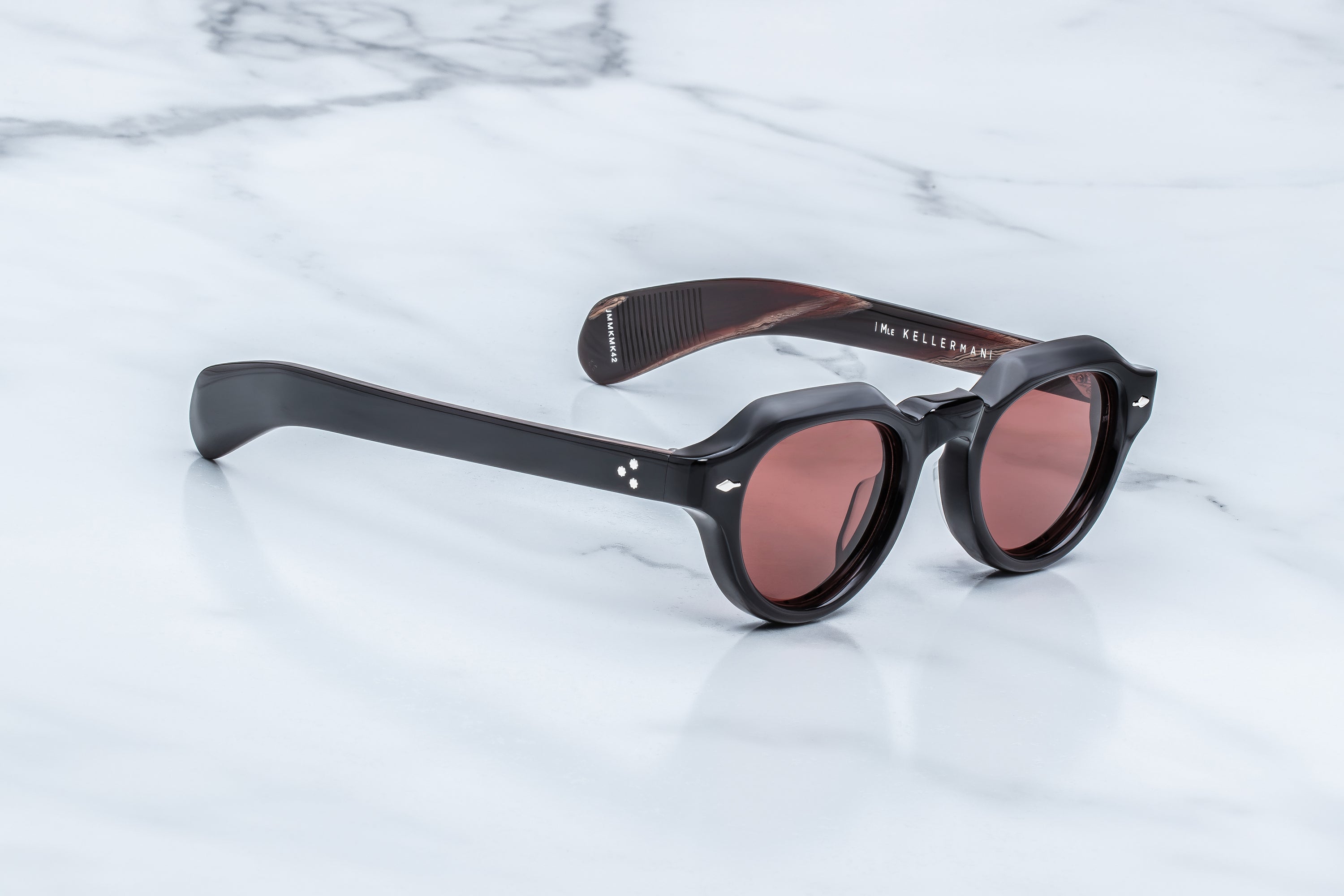 Round sunglasses with thick black frames and pink-tinted lenses, resting on a white marble surface. 