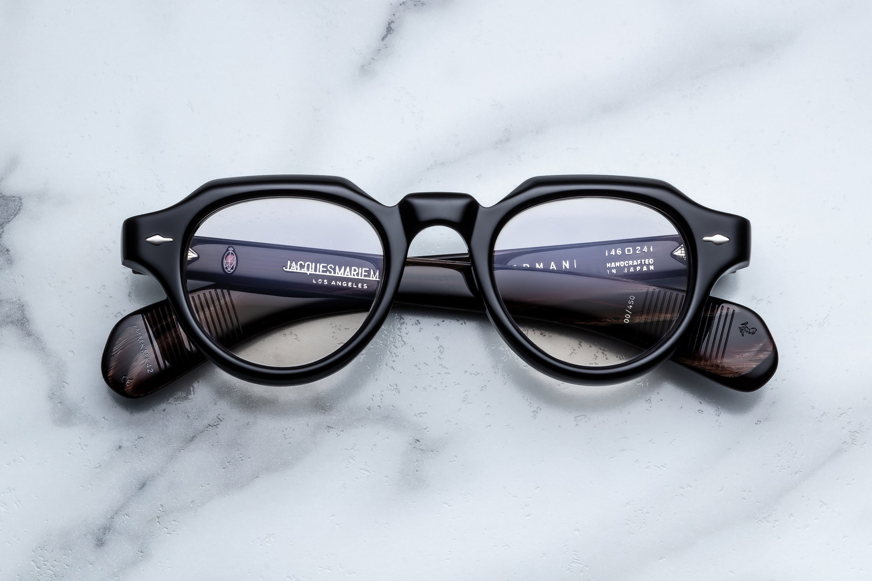 A pair of round-frame eyeglasses with thick black frames and dark brown acetate temples, resting on a white marble surface. 