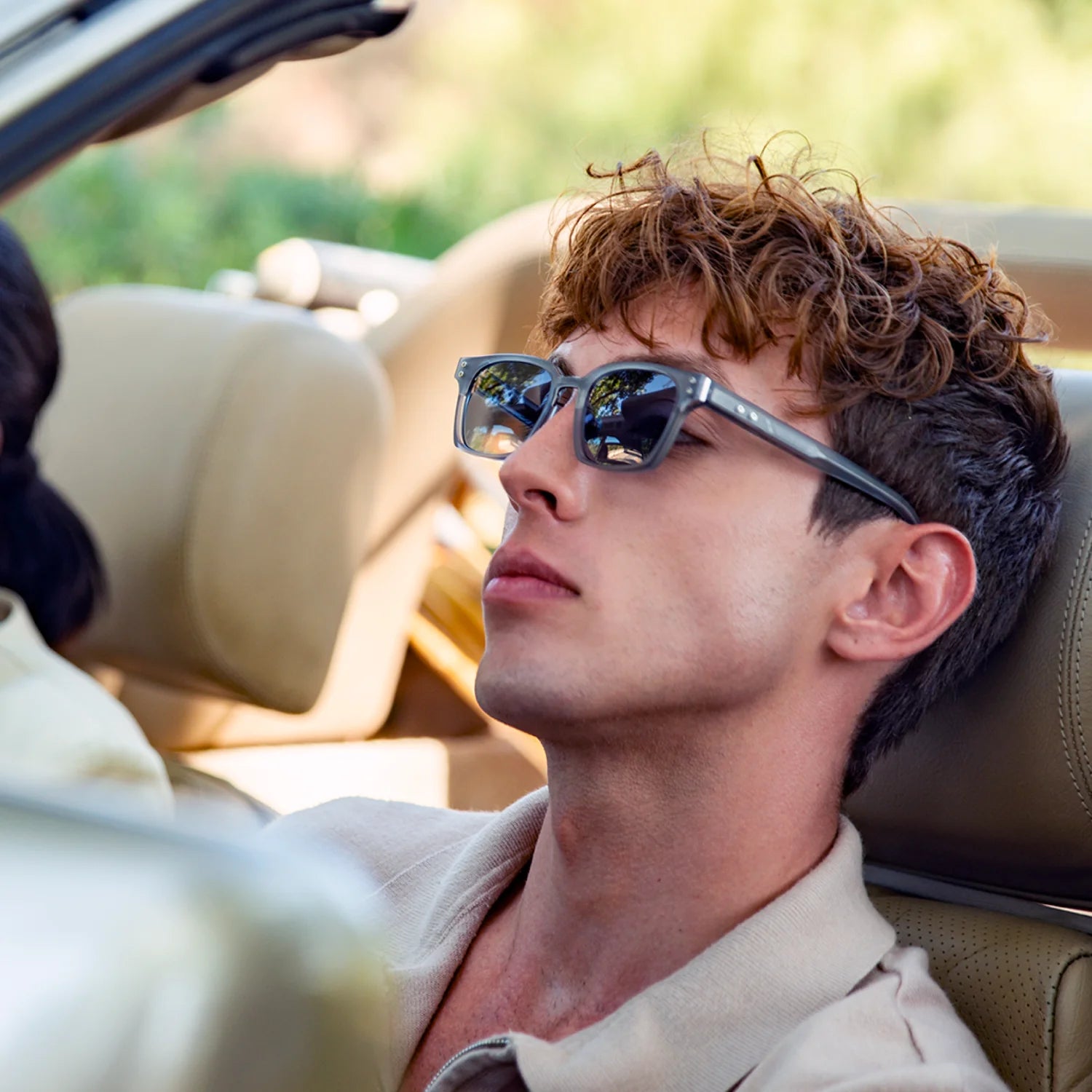 A man with curly brown hair wearing silver-framed sunglasses and a beige shirt, sitting in the passenger seat of a convertible car.