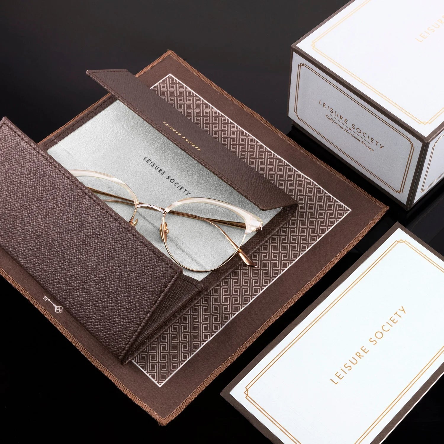 A pair of thin gold-framed eyeglasses resting on a textured brown leather case, which is placed on a patterned brown insert. White and brown LEISURE SOCIETY branded packaging boxes are arranged around it on a dark background.