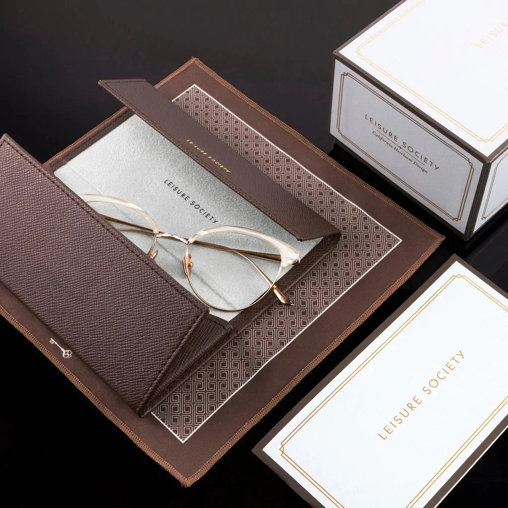 Gold-rimmed, limited edition Leisure Society The Vault: Geiger eyeglasses with photochromic blue tint lenses resting on an open brown leather case, beside a branded box and information card.