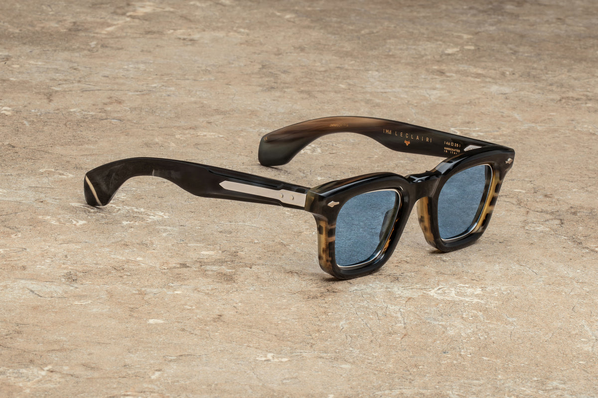 Jacques Marie Mage Heritage 2: Leclair sunglasses by Jacques Marie Mage, featuring a tortoiseshell square frame inspired by classic Art Deco design and blue-tinted lenses, rest on a textured light brown stone surface.