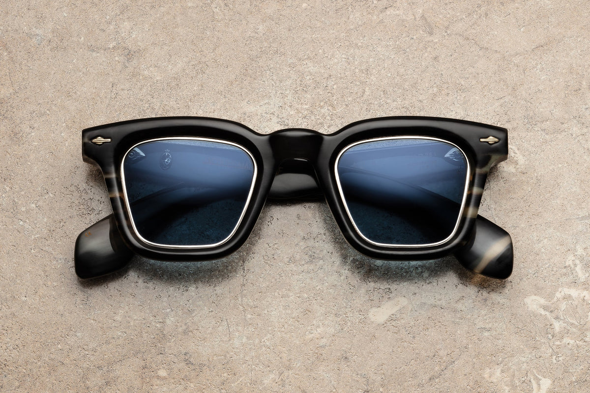 Jacques Marie Mage Heritage 2: Leclair sunglasses by Jacques Marie Mage, featuring black frames and blue-tinted lenses inspired by classic Wellington spectacles, are displayed on a light brown stone surface in an overhead view.
