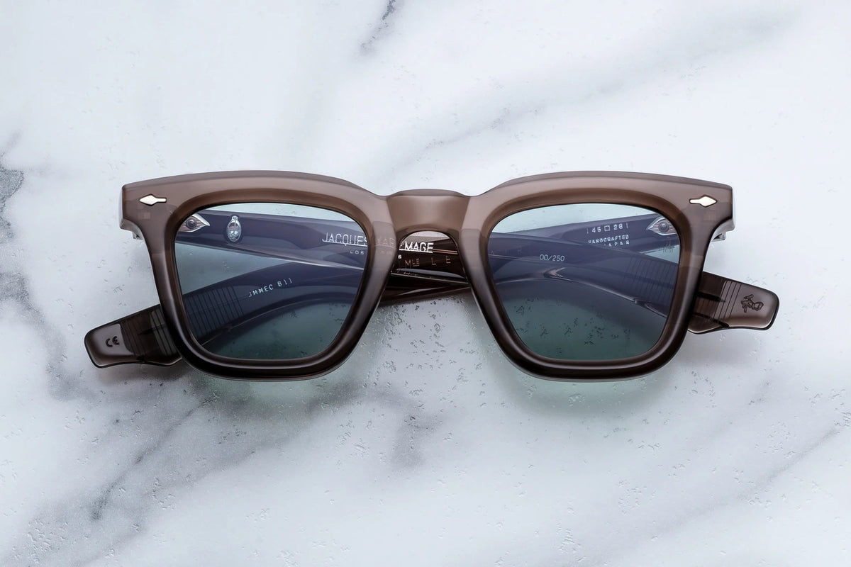 Jacques Marie Mage Leclair translucent brown Wellington-shaped spectacles with silver-tone decorative rivets resting on a marble surface, capturing a classic 50s-inspired silhouette.