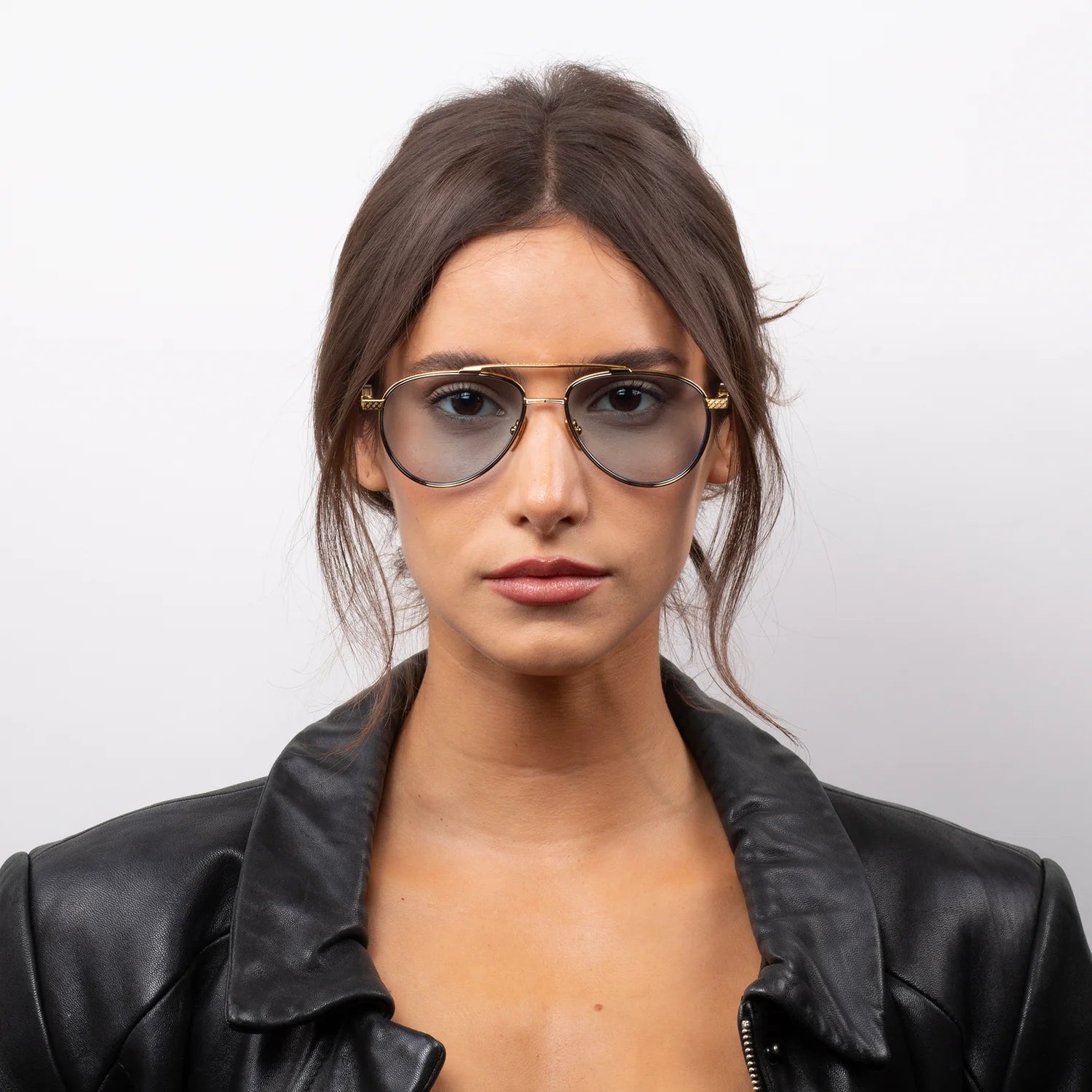 Woman with long brown hair wearing Leisure Society Lucienne luxury eyewear—gold aviator glasses featuring a sleek photochromic lens—paired with a black leather jacket, looking directly at the camera against a plain white background.