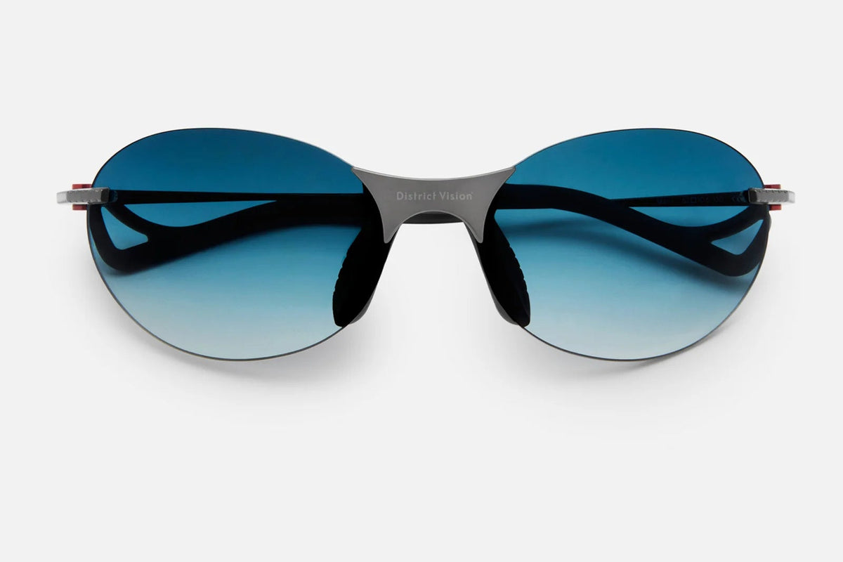 The District Vision Mami Vantage Ti sunglasses by District Vision feature an oval shape, dark blue gradient lenses, and thin black frames with sleek titanium temples for enhanced durability and style, displayed on a plain light grey background.